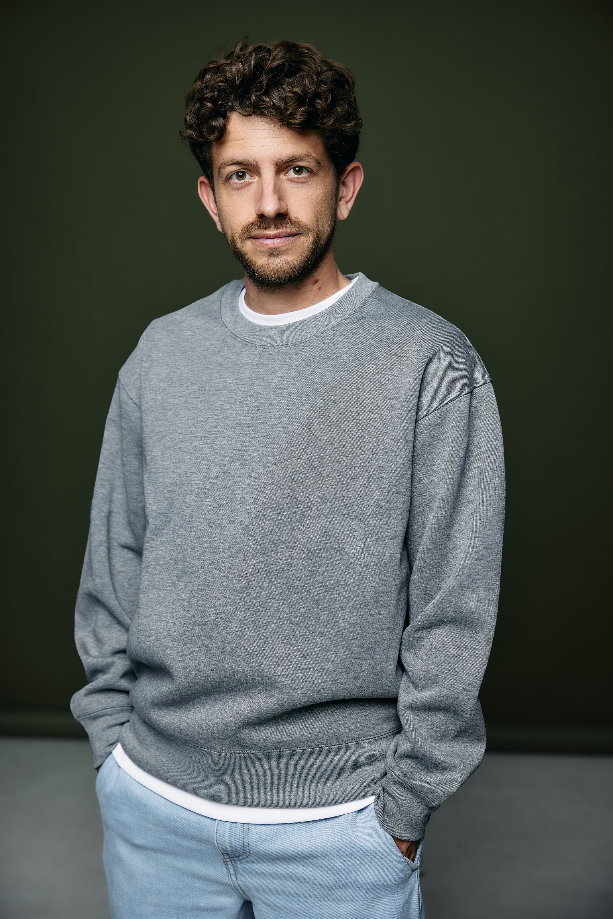 A man with curly dark hair and a beard, wearing a gray sweatshirt and light blue jeans, standing against a dark green background.