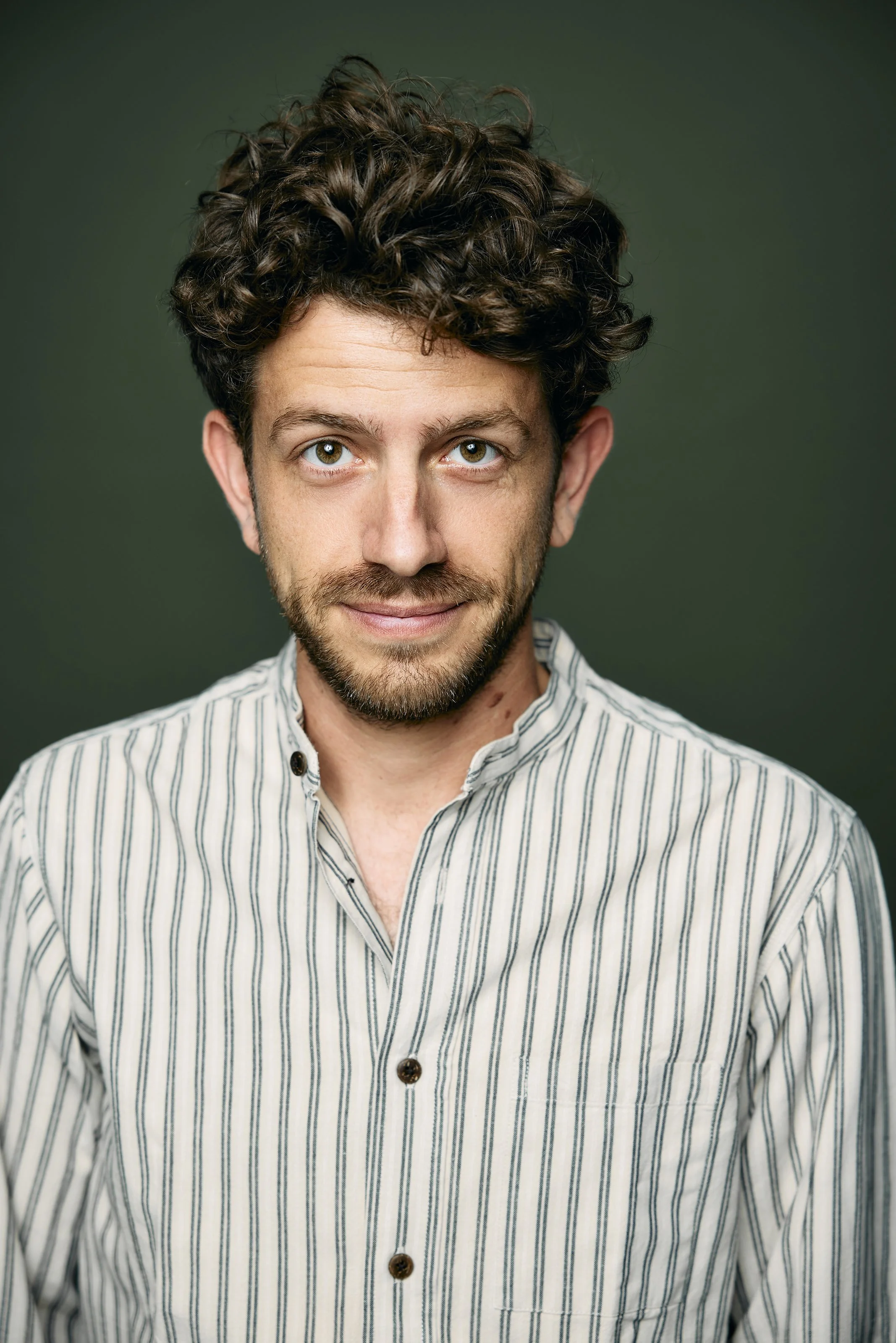 A man with curly brown hair, blue eyes, and a beard, wearing a striped shirt, smiling at the camera against a green background.