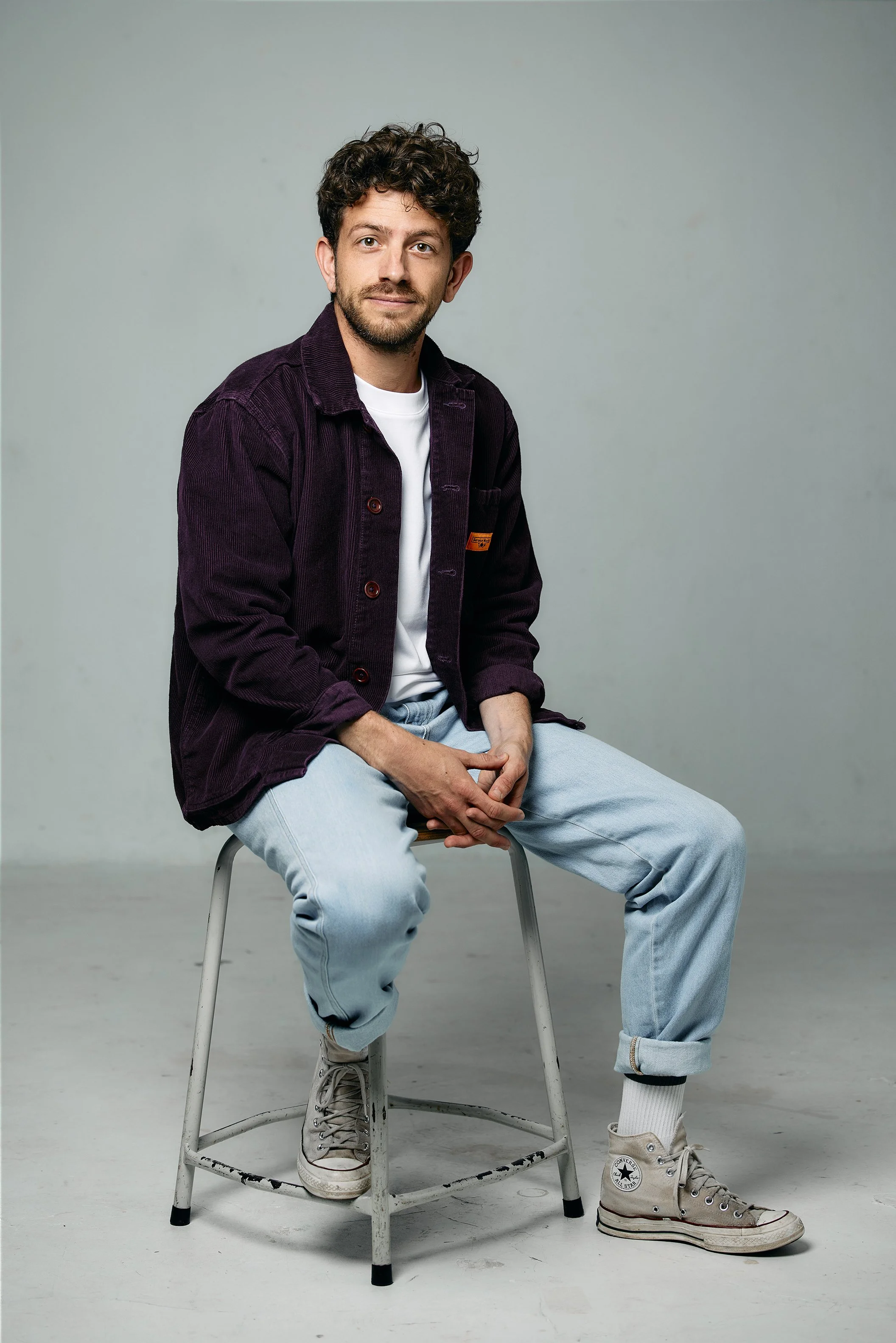 A young man with curly hair, beard, wearing a burgundy jacket, white t-shirt, light blue jeans, and gray high-top Converse sneakers, sitting on a metal stool against a plain light gray background.