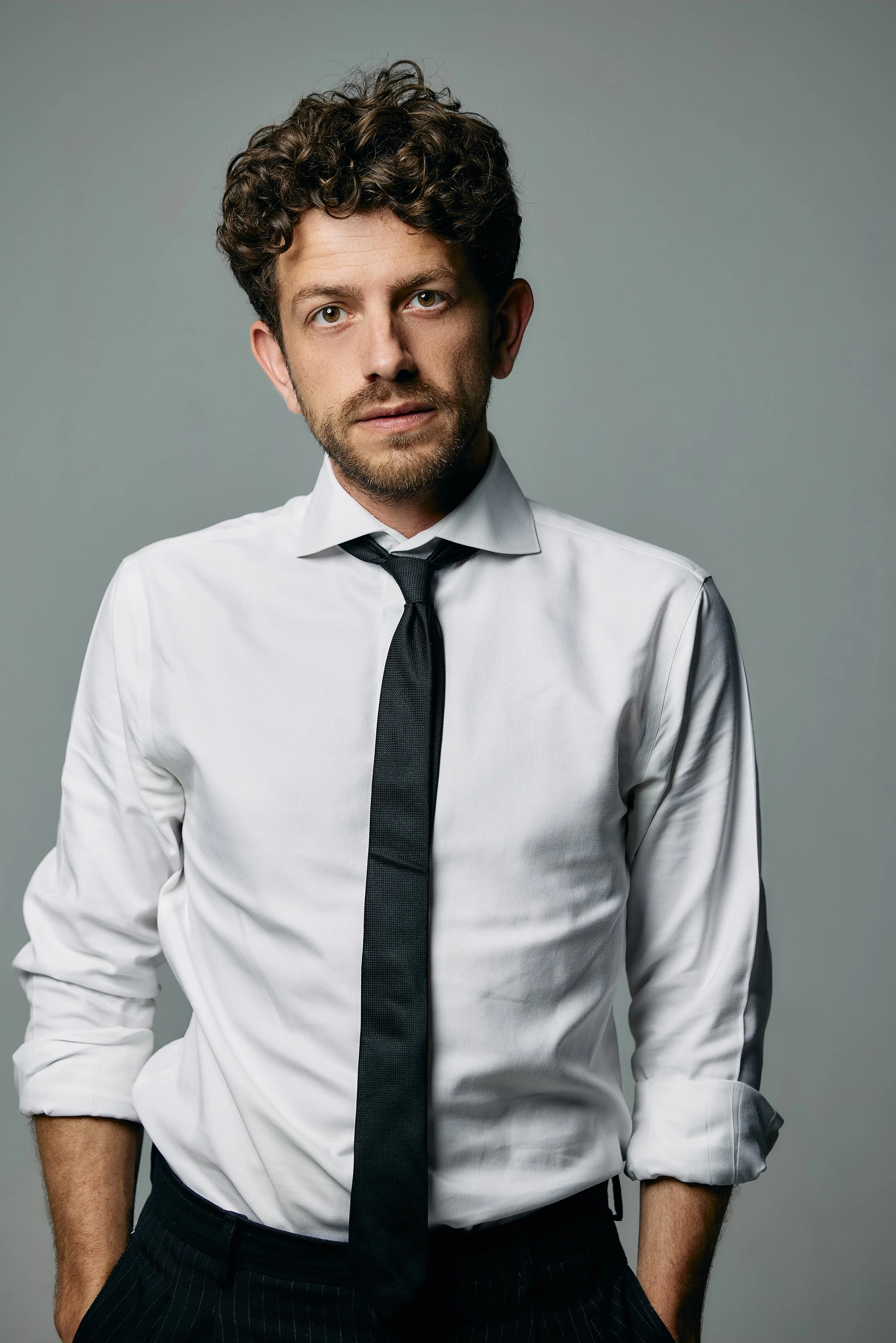 Portrait of a man with curly brown hair, a beard, wearing a white dress shirt with sleeves rolled up, and a black tie, standing against a gray background.