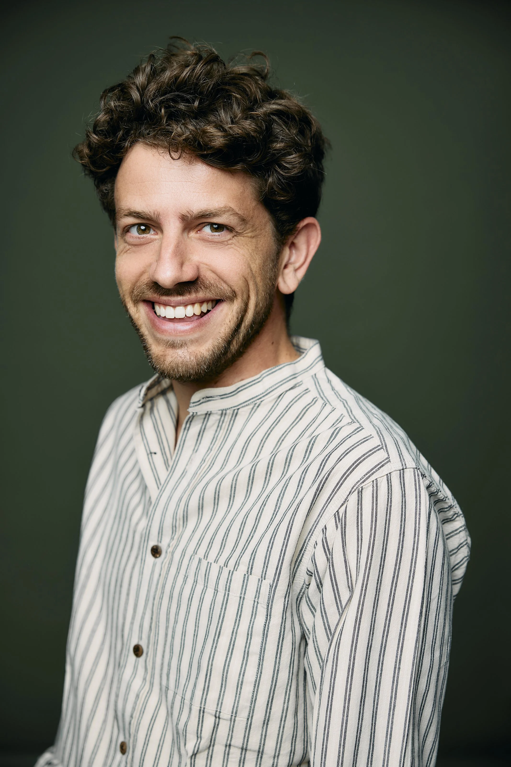 A smiling man with curly brown hair, light beard, wearing a striped button-up shirt, against a dark green background.