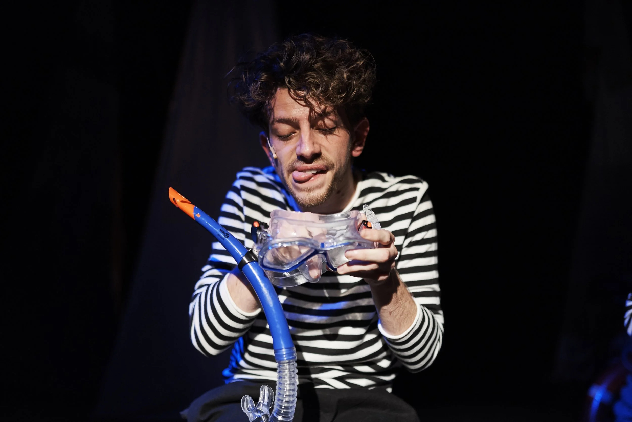A man with curly hair in a striped shirt is holding and looking at a snorkel mask with a snorkel attached, on a dark background.