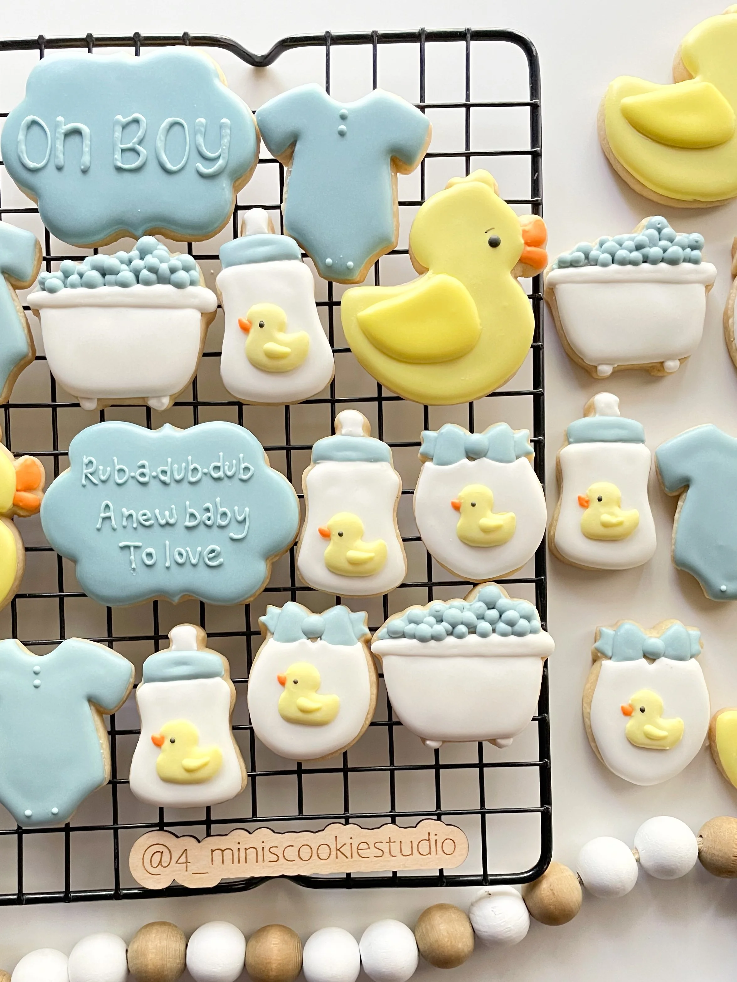 Baby shower cookies on a black wire rack, decorated with pastel blue, yellow, and white icing. Cookies include words, baby bottles, ducks, onesies, and bathtubs with bubbles.