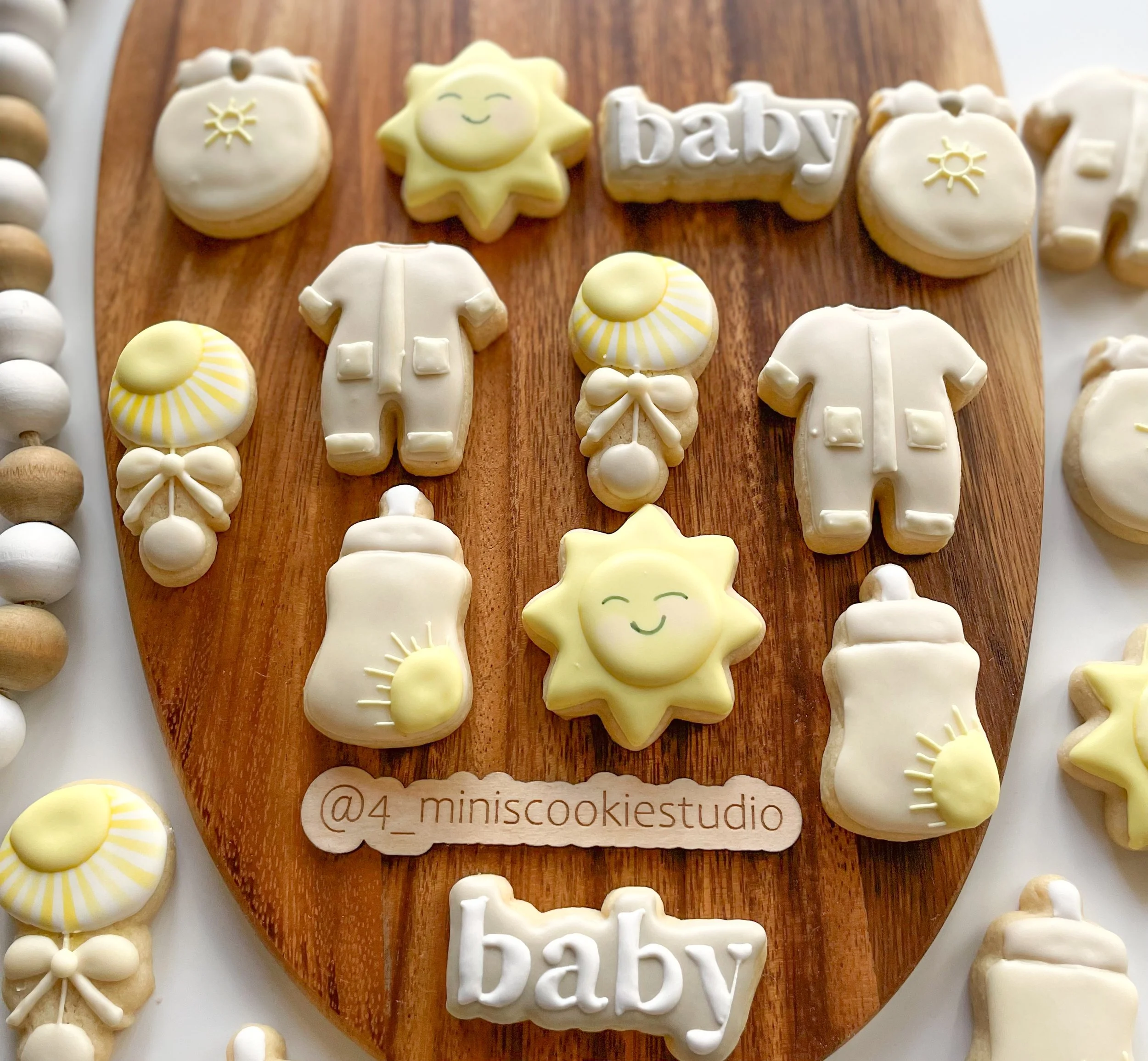 Assorted baby-themed cookies on a wooden board, including ones shaped like baby onesies, bottles, suns, and pacifiers with pastel colors and cheerful smiling faces, surrounded by additional cookies on a white surface.