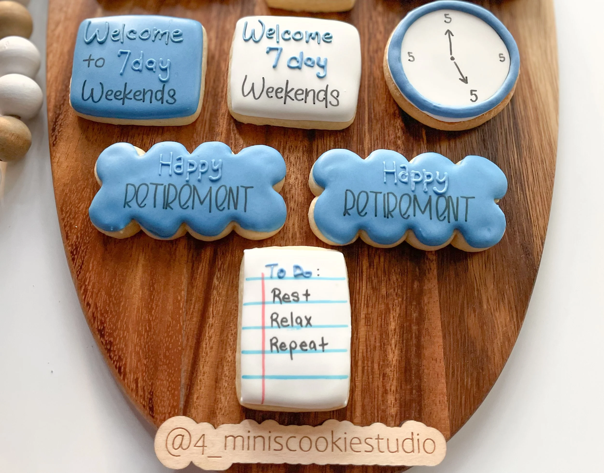 Decorative cookies on a wooden board with messages about retirement and weekends, including two cloud-shaped cookies that say "Happy Retirement," a rectangular cookie with a to-do list, a clock cookie, and two square cookies that say "Welcome to 7 da