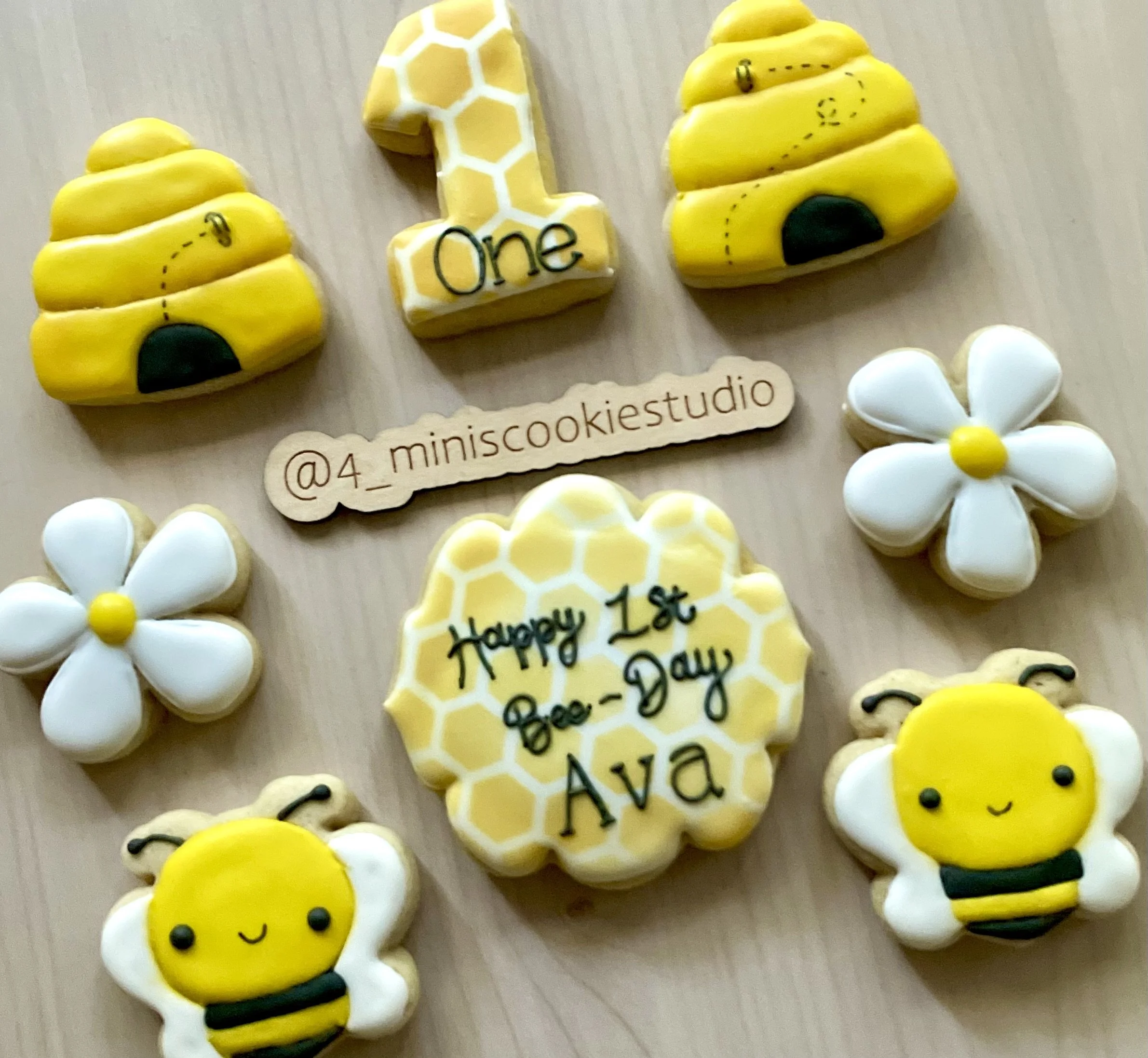 Decorated cookies celebrating a first bee day, including bee, honeycomb, and flower shapes, arranged on a light wooden surface.