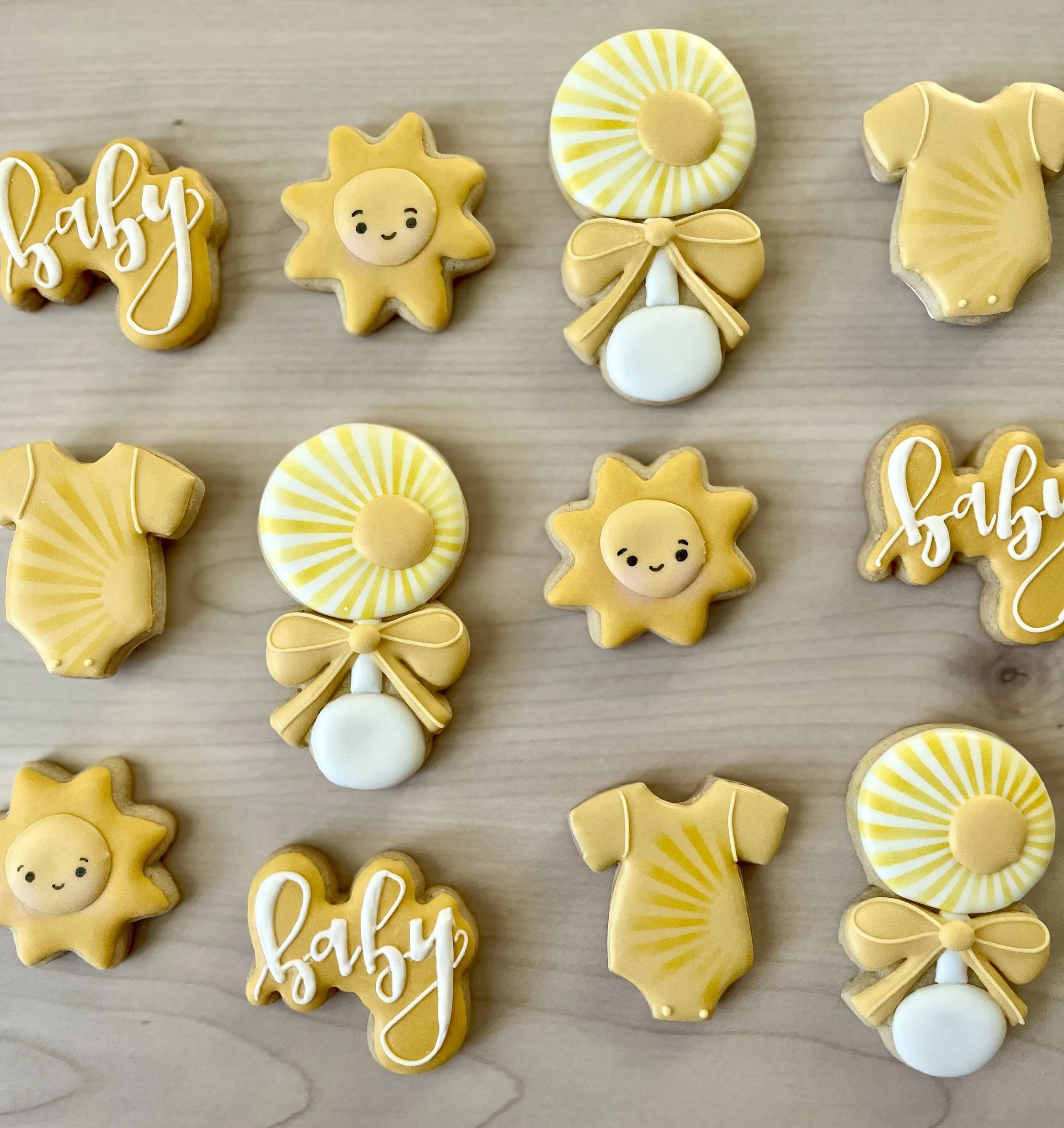 Collection of yellow-themed baby shower cookies on a wooden surface, including sun designs, baby onesies, and baby bottles with bows.