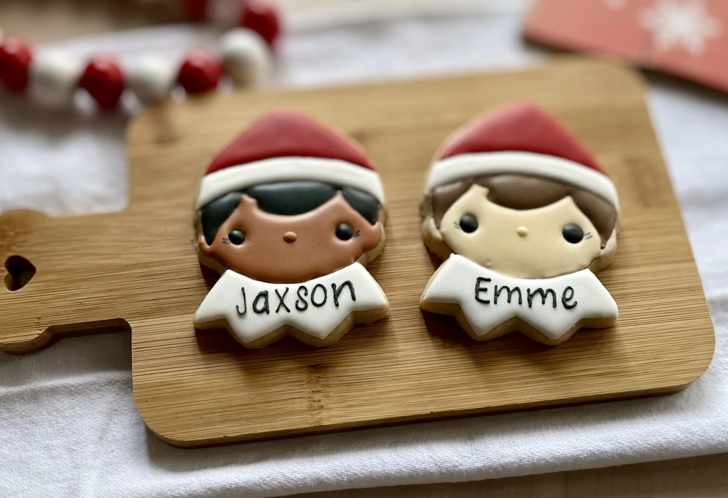 Cookies decorated as children wearing Santa hats, labeled with the names Jaxson and Emme, on a wooden board.