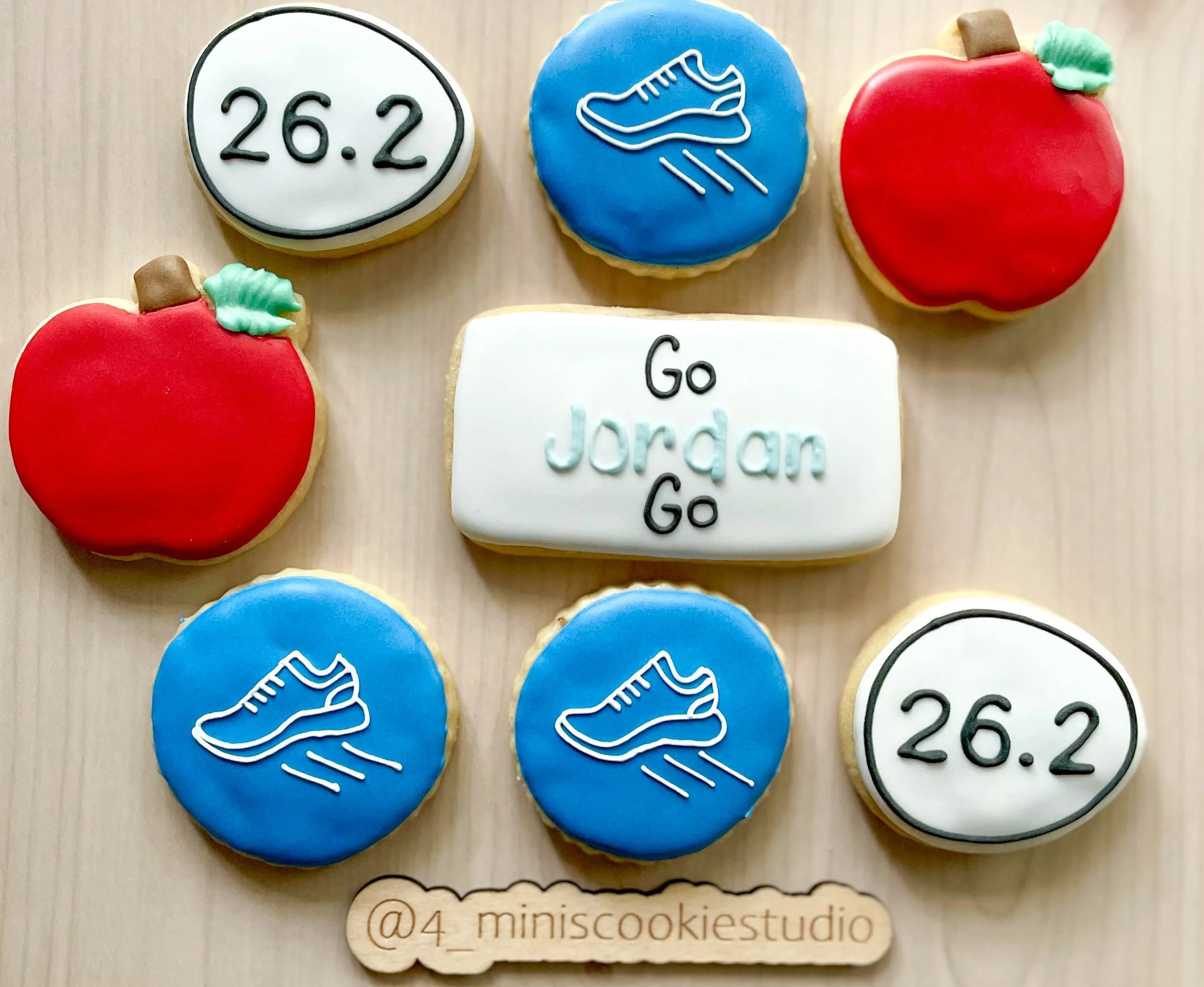 A collection of decorated cookies themed around running and a motivational message. There are two red apple cookies, two blue shoe cookies, two white cookies with the number 26.2, and a rectangular white cookie with the text "Go Jordan Go". The cooki