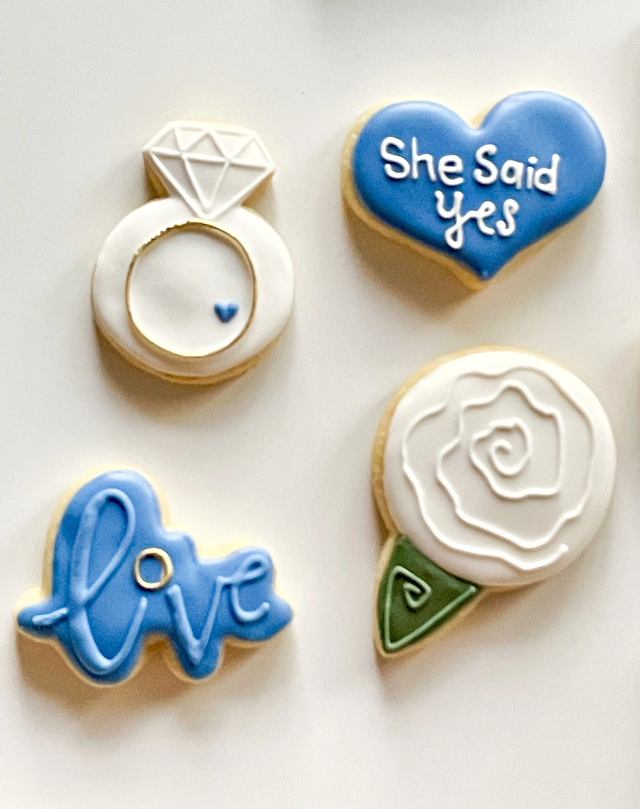 Decorative cookies with messages: a diamond ring, a heart with the text 'She Said Yes', a cookie with the word 'love', and a cookie shaped like a flower with a green leaf.
