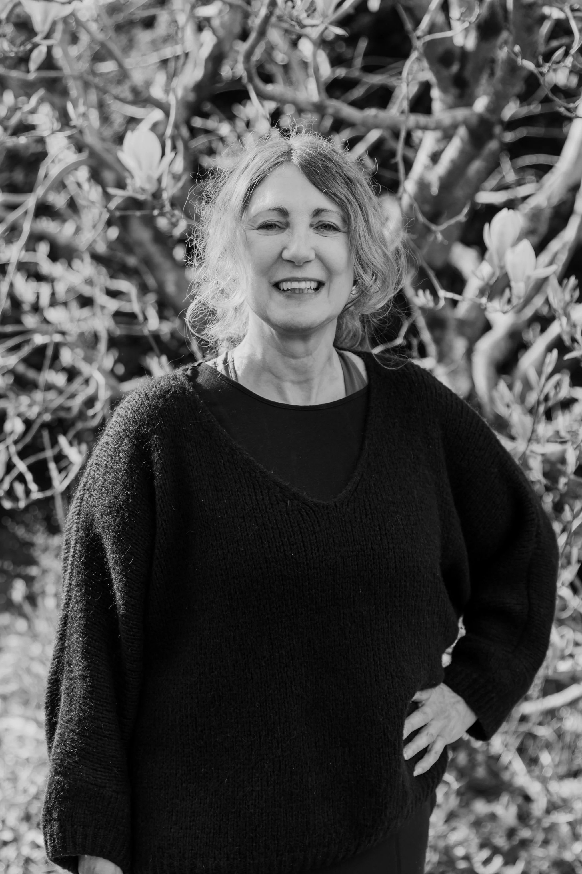 A woman smiling outdoors, standing in front of a background of foliage and tree branches.