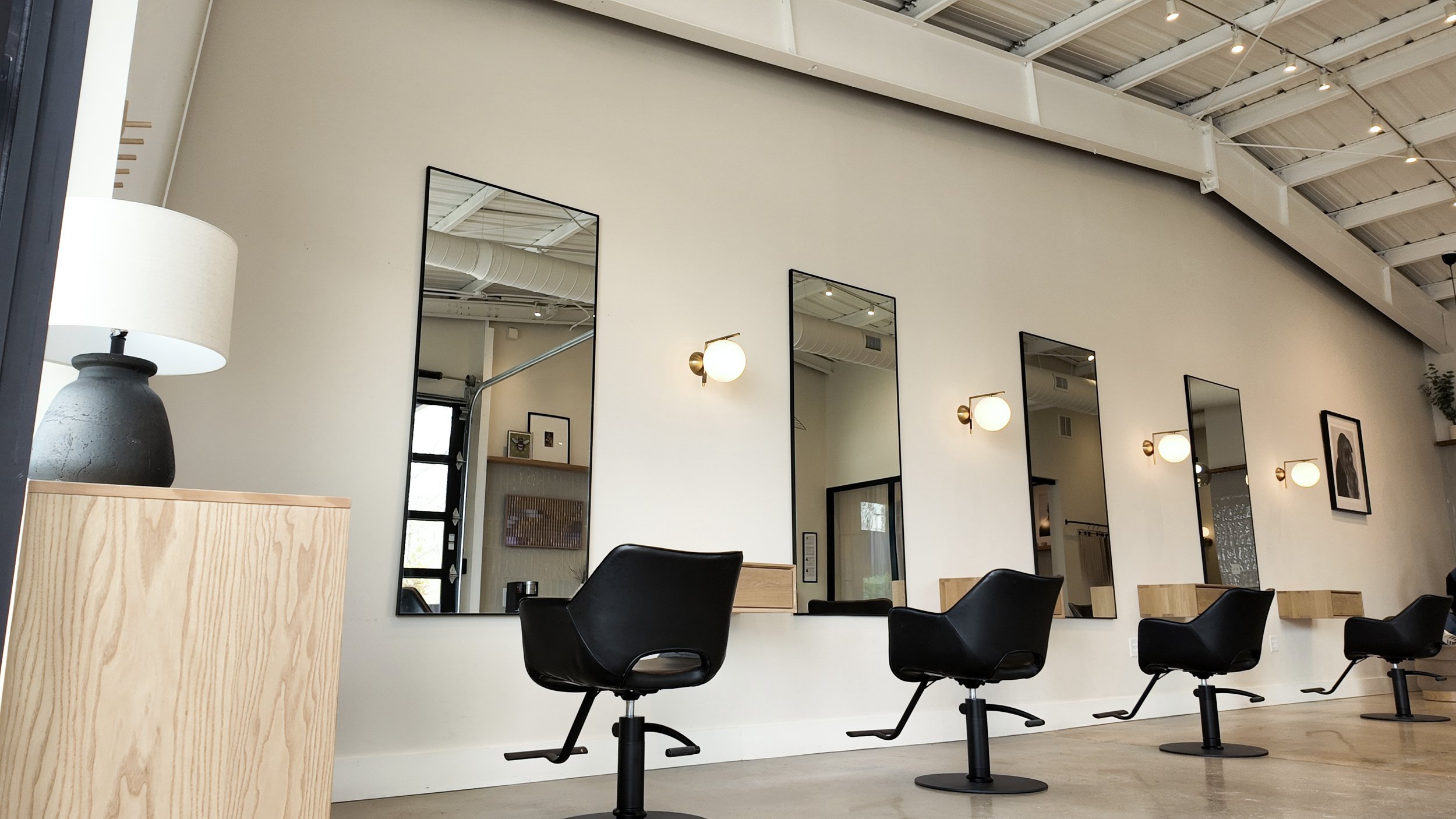 Empty hair salon with four black styling chairs in front of tall mirrors, modern wall sconces, framed artwork, and minimal decor.