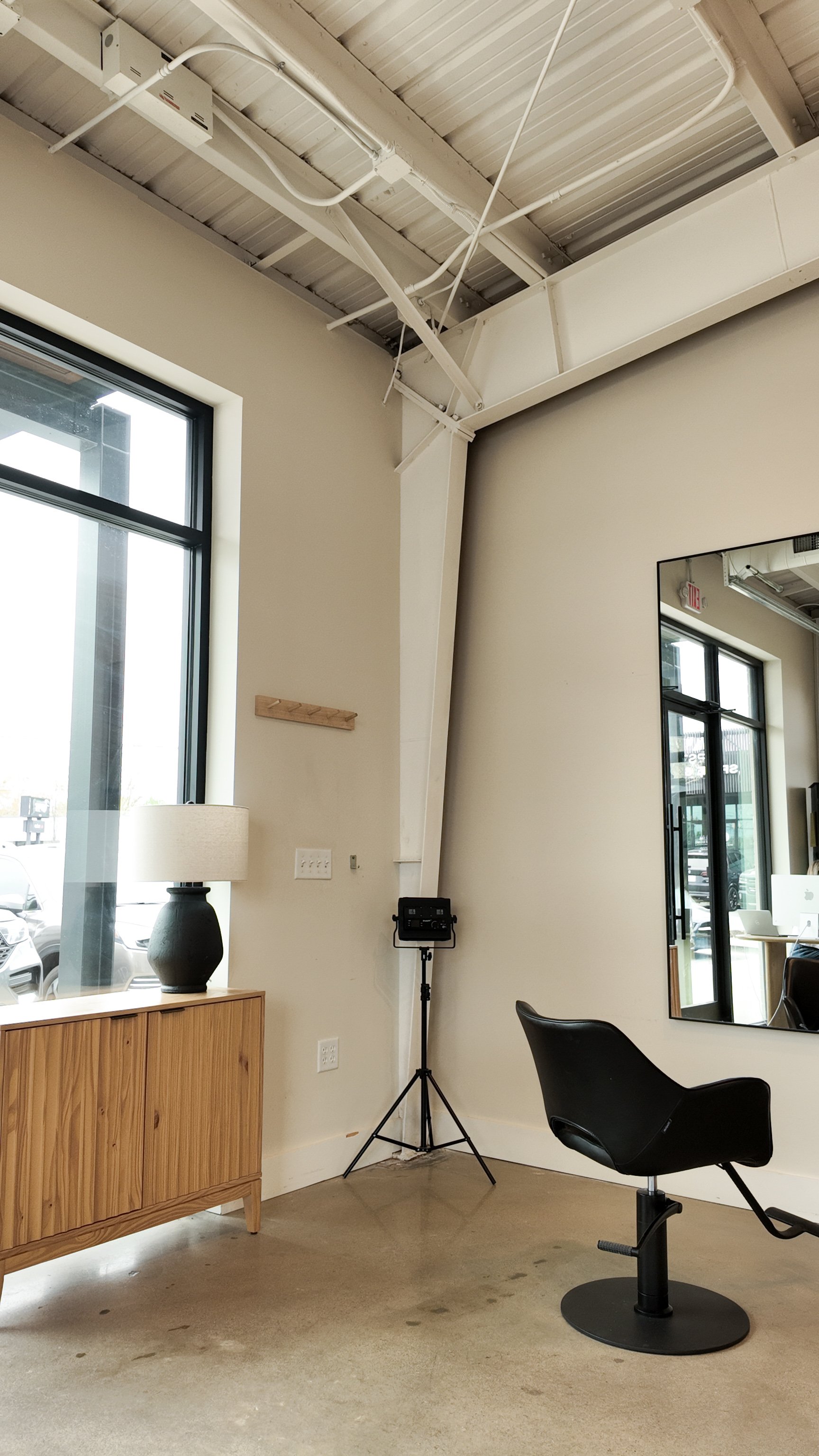 Interior of a minimalistic room with a window, a black salon chair, a wooden sideboard with a lamp, a large mirror, and some equipment in the corner.