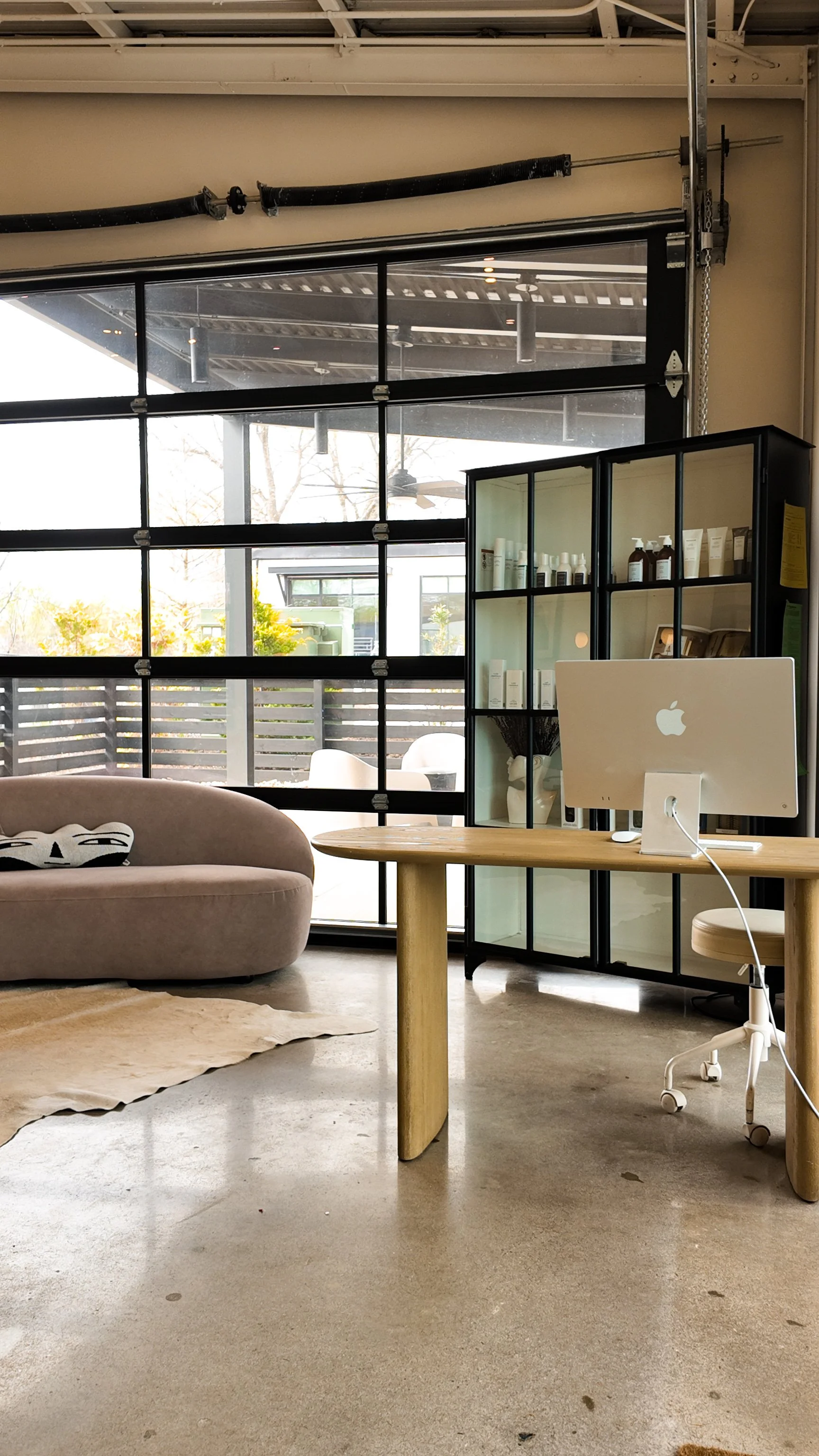 Modern interior workspace with a beige sofa, a wooden table holding an Apple computer, a black glass display shelf with skincare products, and large windows showing an outdoor patio area.