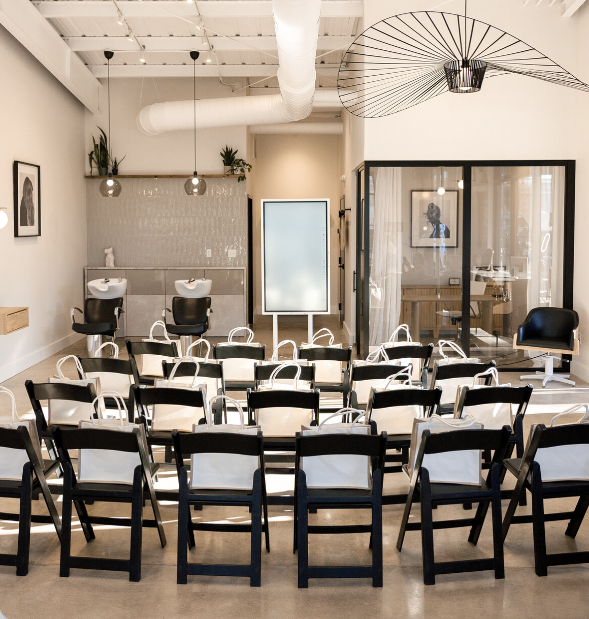Inside a modern salon with chairs arranged for a presentation or class, two washbasins at the back, and a private room with glass walls on the right.