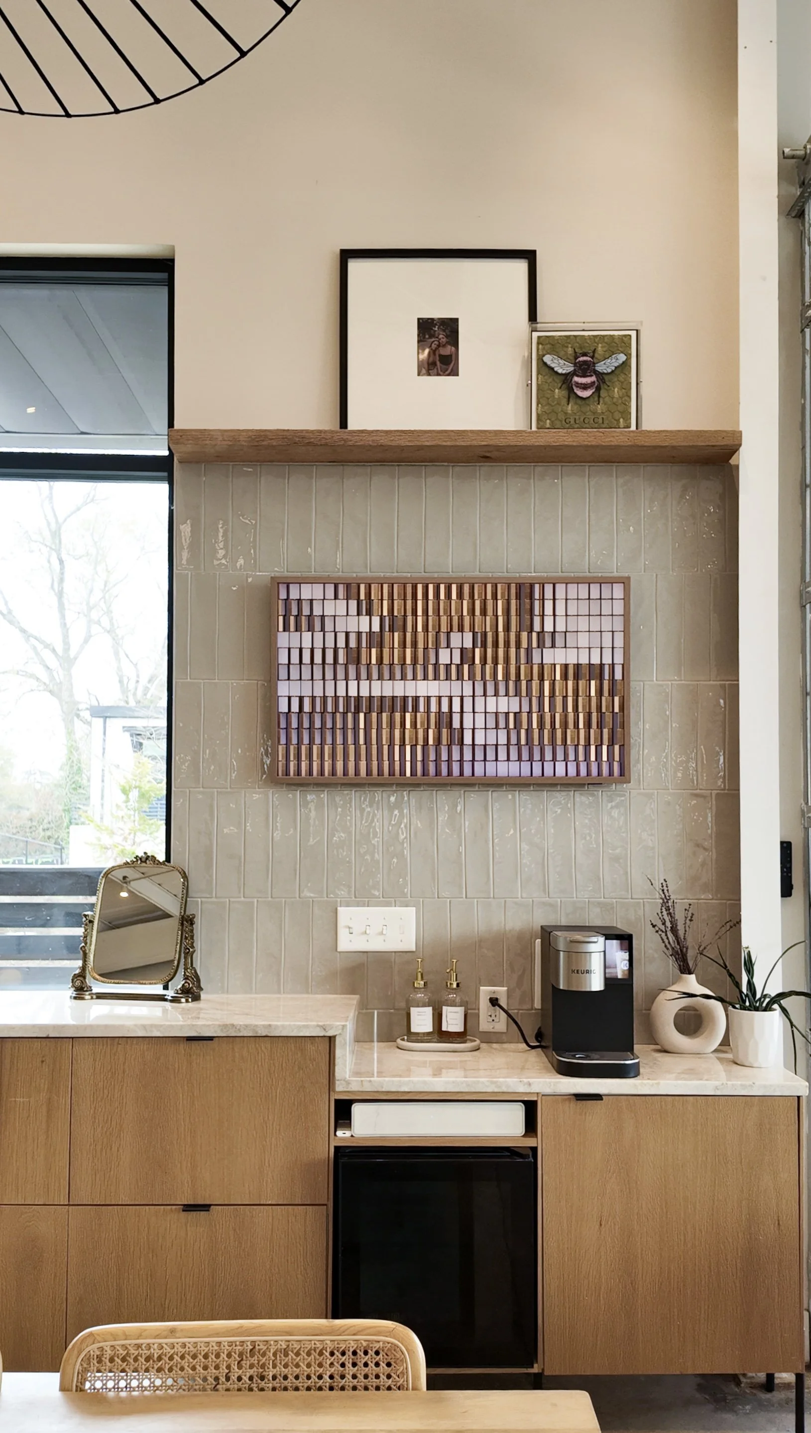 Decorative wall with framed art, a wooden shelf, and a backsplash. Kitchen counter with small decorative mirror, bottles, coffee maker, and vases with plants.