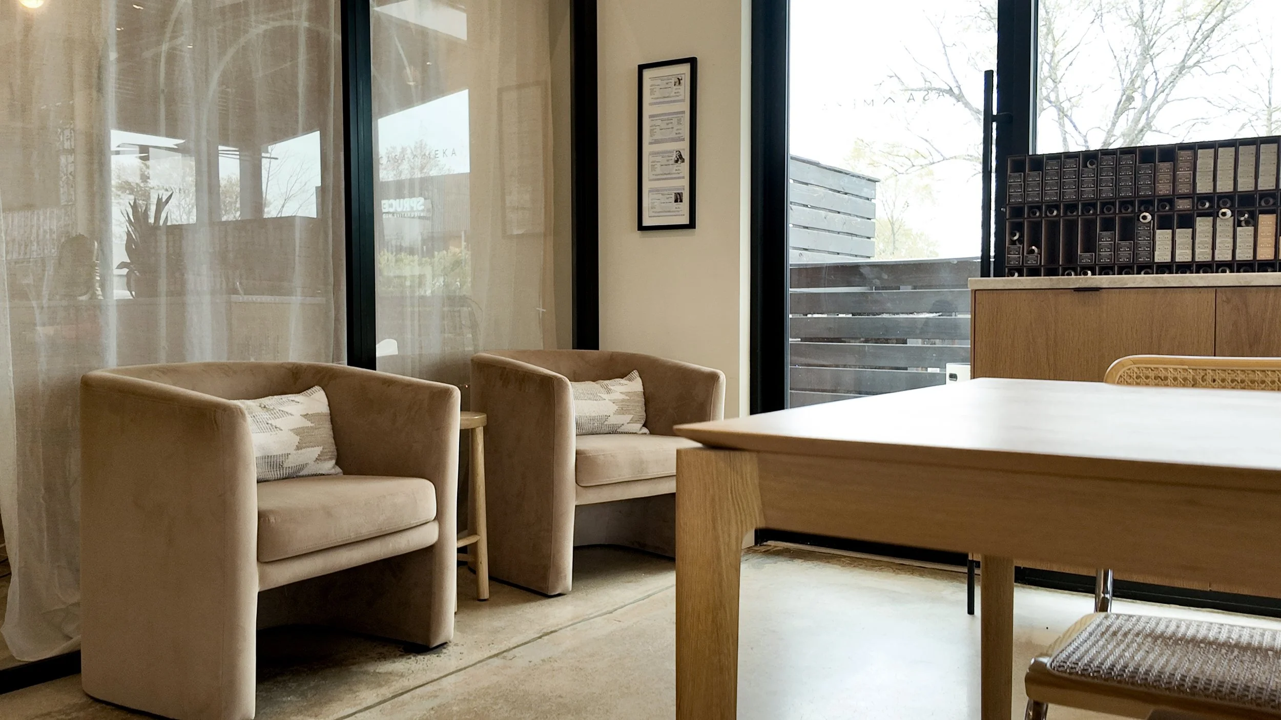 Part of a cozy waiting area with two beige armchairs with pillows, a wooden table and a chair, large glass windows showing trees outside, and a wooden cabinet with small compartments.