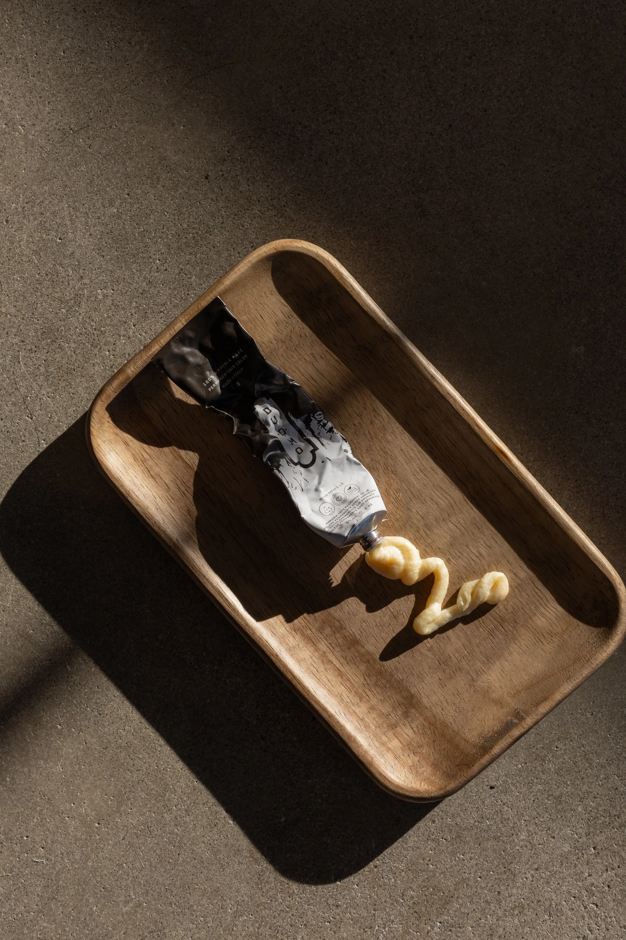 A wooden tray on a gray surface holding a tube of black and white hair color and a twisted yellow paint worm extending from the tube.