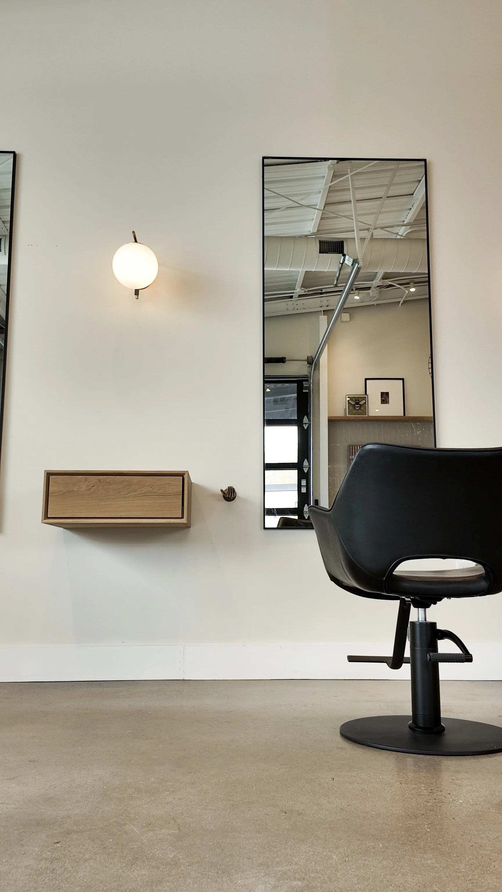 Empty black salon chair in front of a wall with a rectangular mirror, a wall-mounted light, and a small wooden shelf in a minimalistic salon.