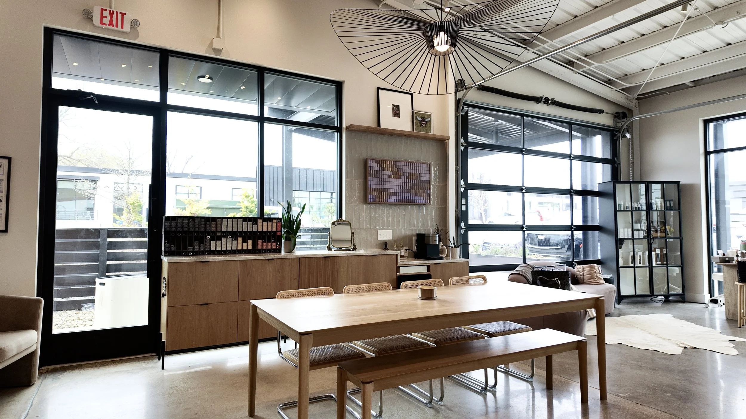 Interior of a modern cafe with large black-framed windows, a wooden dining table with six chairs, a bench, a sideboard with files, and a cozy seating area with a sofa and display cabinet.