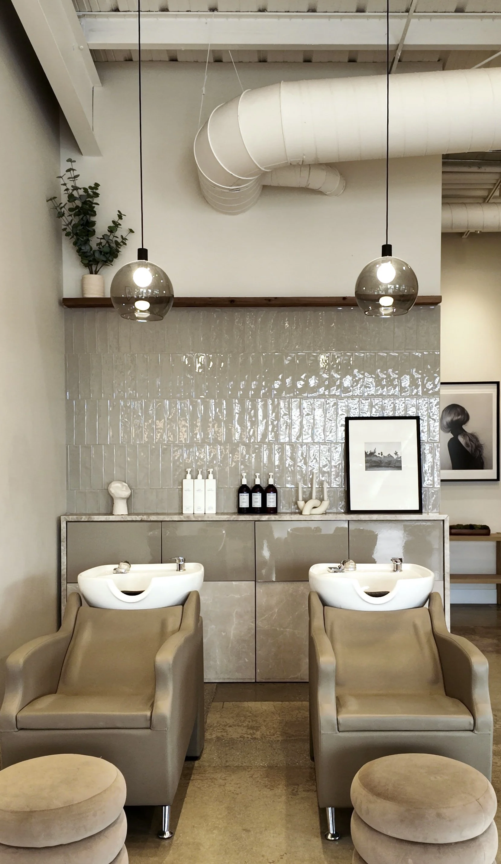 Two beige salon chairs with built-in sinks for hair washing, set in a modern, minimalist hair salon with neutral tones and decorative art.