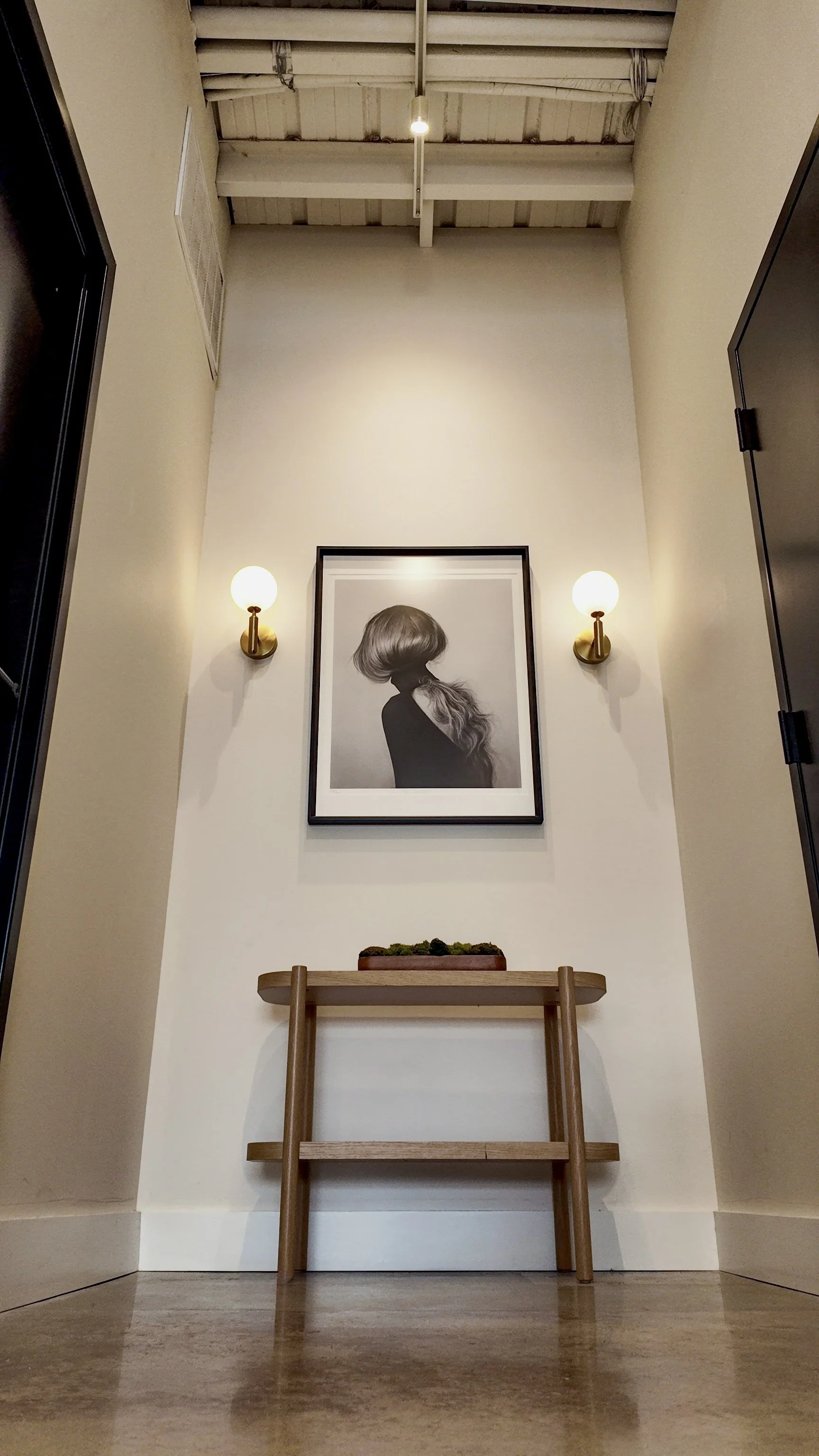 A hallway with a framed black and white portrait of a woman with long hair, two wall sconces with globe lights on either side, a small wooden bench with a rectangular planter on top, and an exposed ceiling with lighting fixtures.