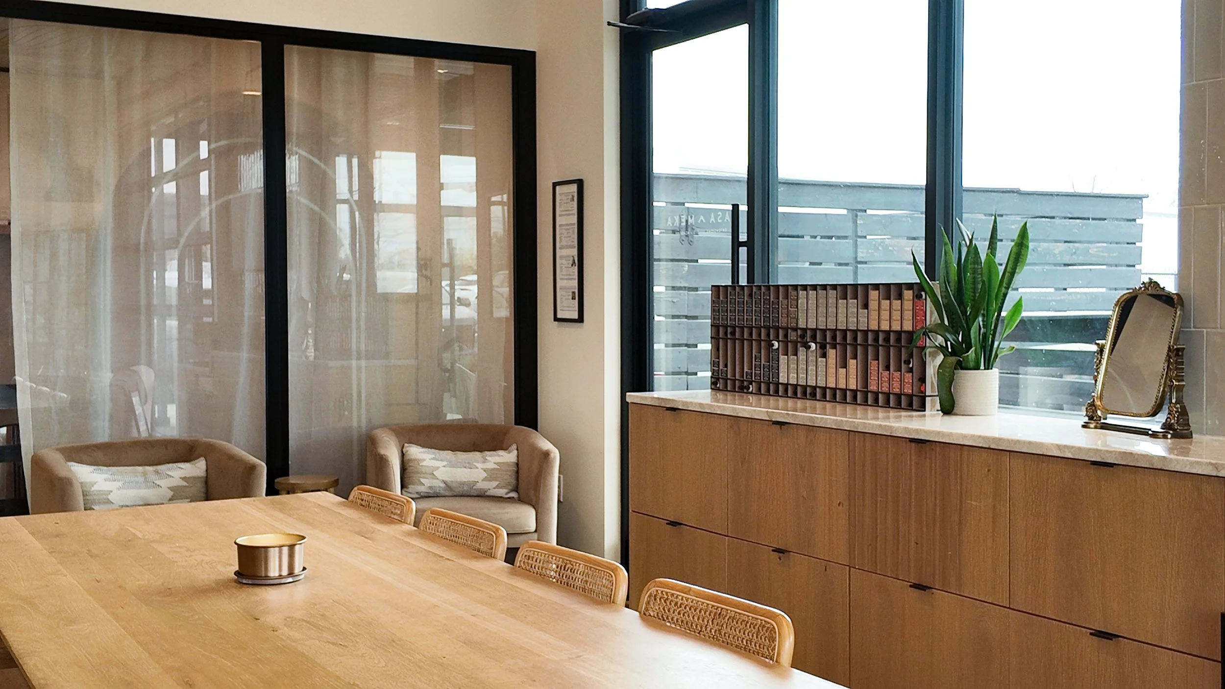 Modern interior with a wooden table and chairs, beige armchairs with patterned cushions, a wooden sideboard with a potted plant, a small mirror, and a window with a wooden privacy screen.