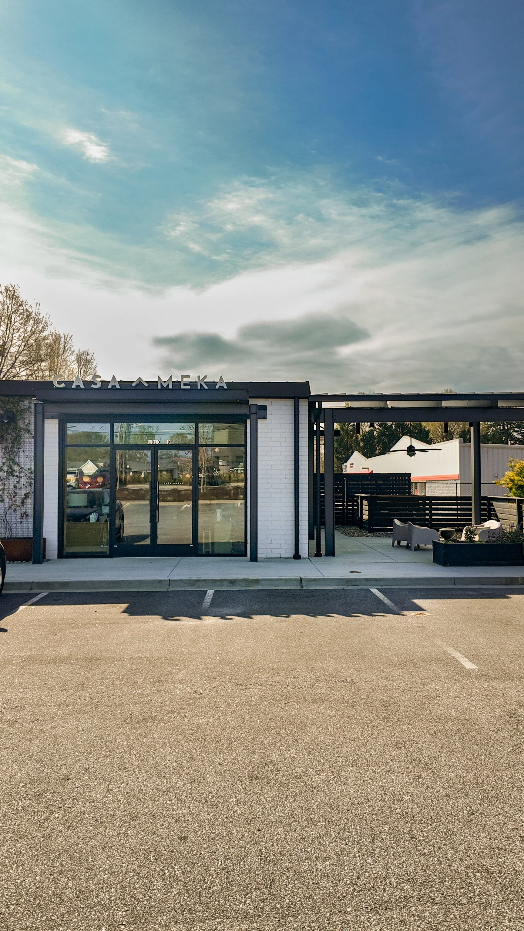 Front view of a modern building with a sign that reads 'Casa Meka', parking lot in front, and an outdoor seating area with chairs and a ceiling fan.