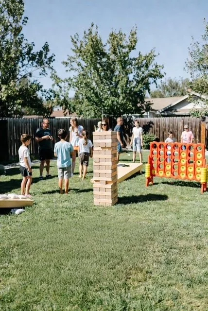 Children playing outdoors in a backyard with a giant Jenga game and a giant Connect Four game on the grass, surrounded by a fence and trees.