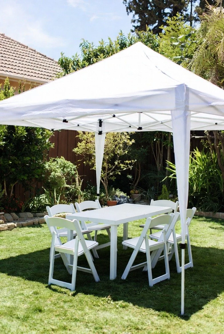 A white outdoor canopy tent with six white chairs surrounding a white table in a backyard with green grass and trees.