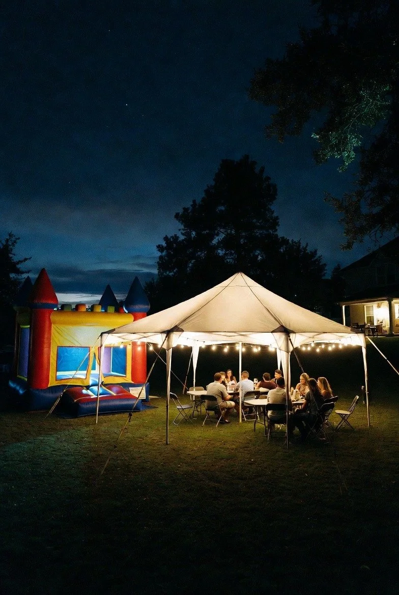 People gathered around at night under a lit canopy tent with string lights, adjacent to a colorful inflatable bounce house on a grassy yard, with trees and a house in the background.