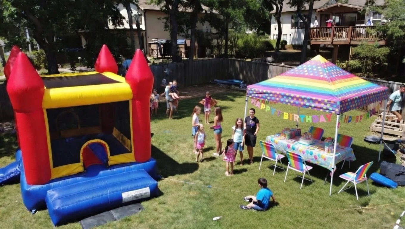 Children and adults gather in a backyard on a sunny day for a birthday party, with a colorful bounce house, a decorated table under a rainbow-colored canopy, and a birthday banner reading 'Happy Birthday'.