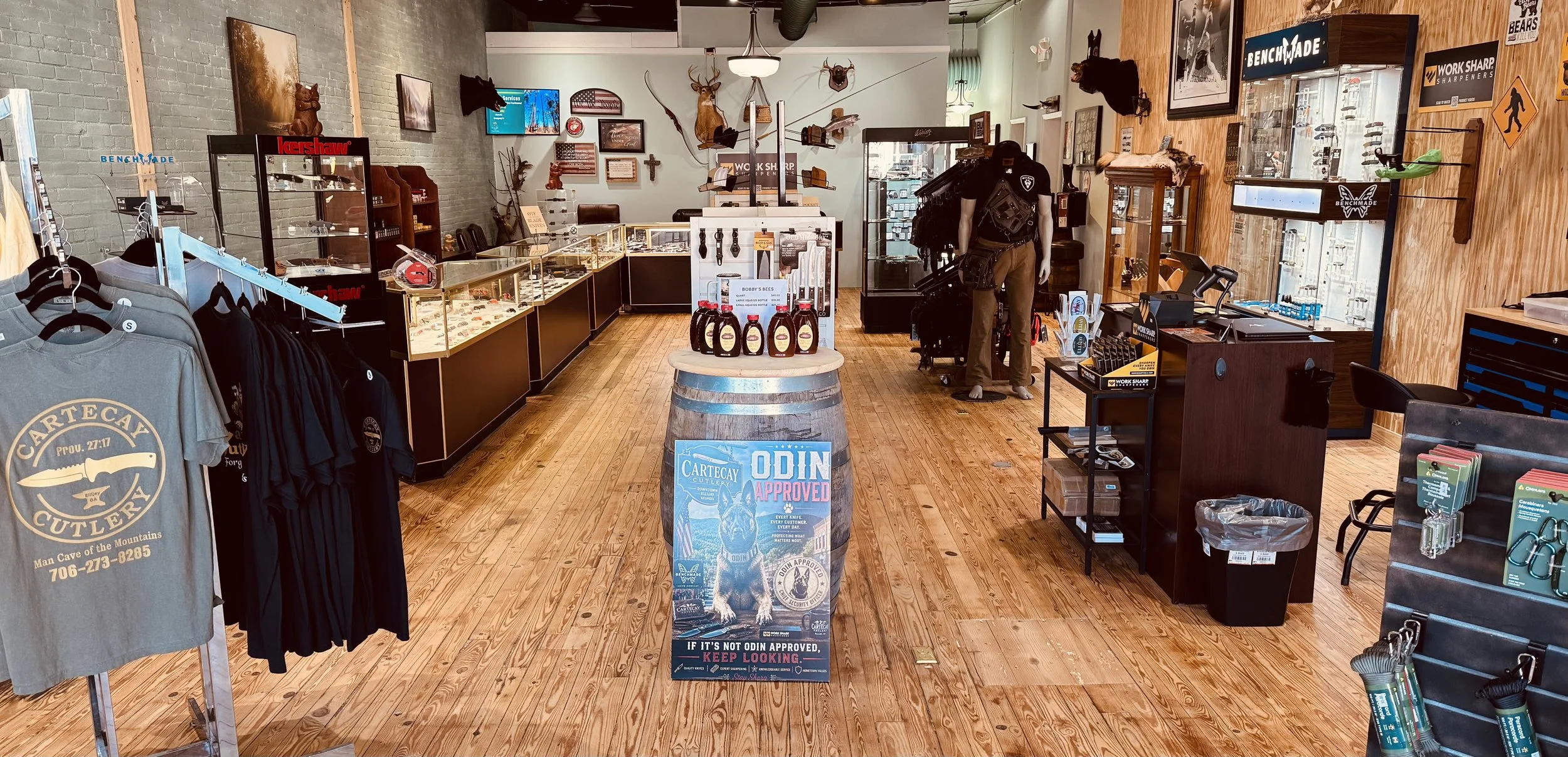 Interior of a retail store selling outdoor gear, knives, and accessories, featuring wooden floors, display counters, clothing racks, taxidermy animals, and various signs and decorations on the walls.