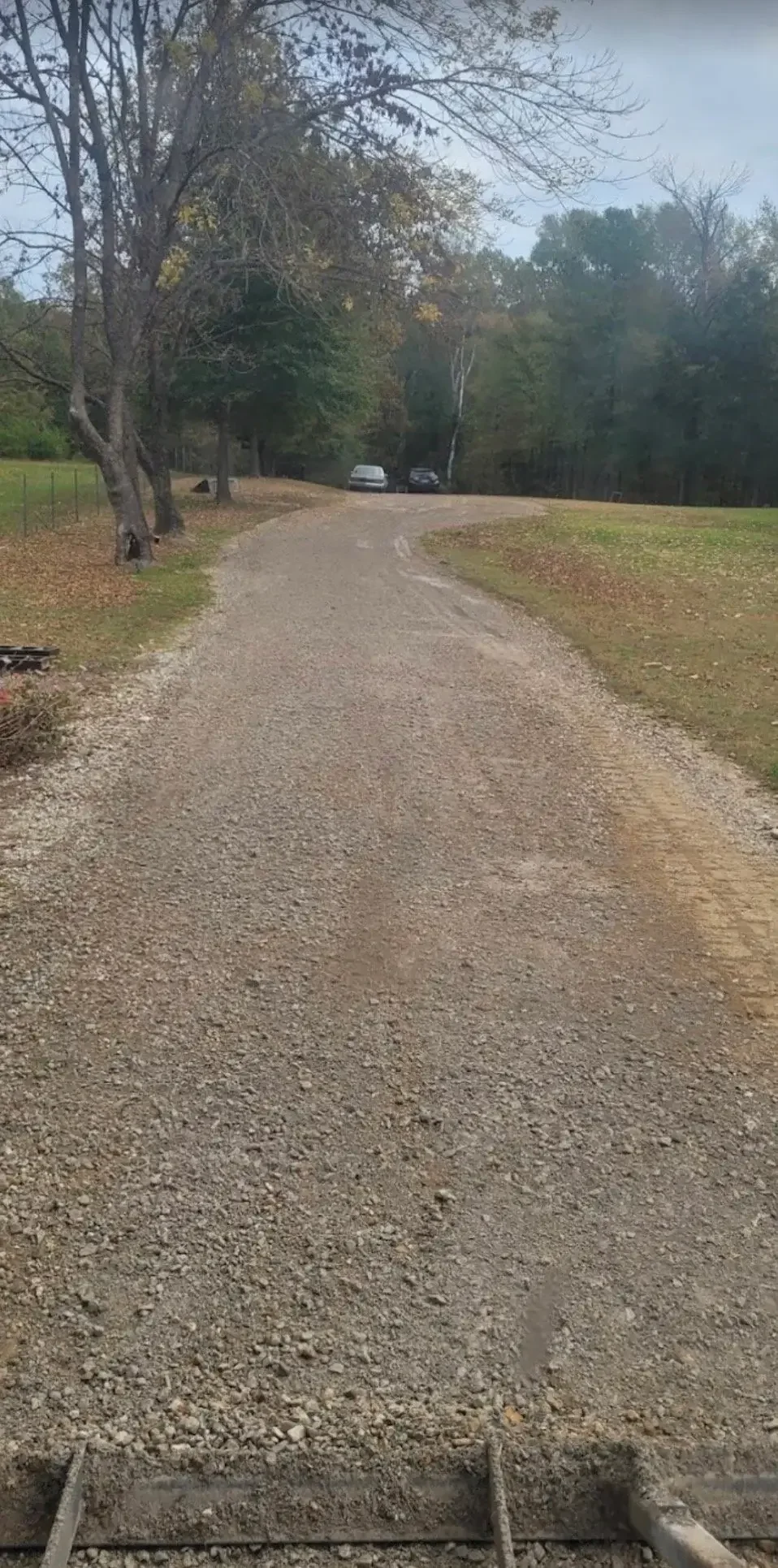 A gravel driveway with two parked cars and trees on either side, leading into a wooded area with a cloudy sky overhead.