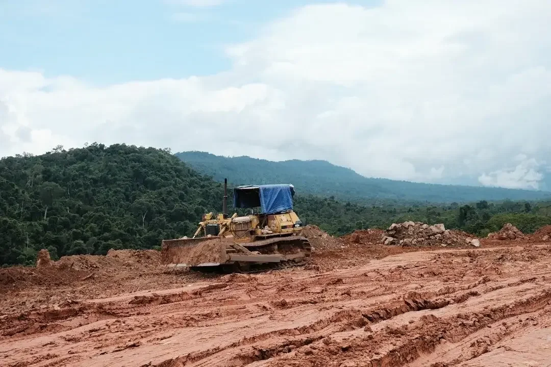 A bulldozer working on a construction site with dirt and mud, with lush green hills and mountains in the background under a partly cloudy sky.