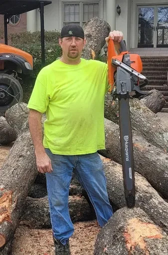 Man in a bright yellow shirt and black cap holding an orange and black chainsaw, standing in front of a pile of chopped logs outside a house.