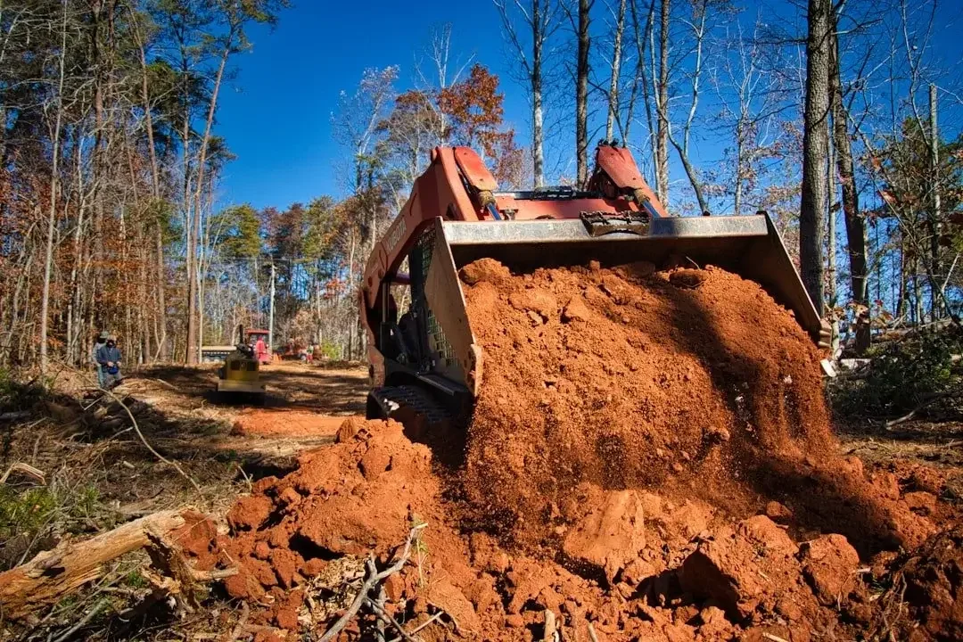 Excavator moving earth on a construction site in a forested area with trees and a clear blue sky.