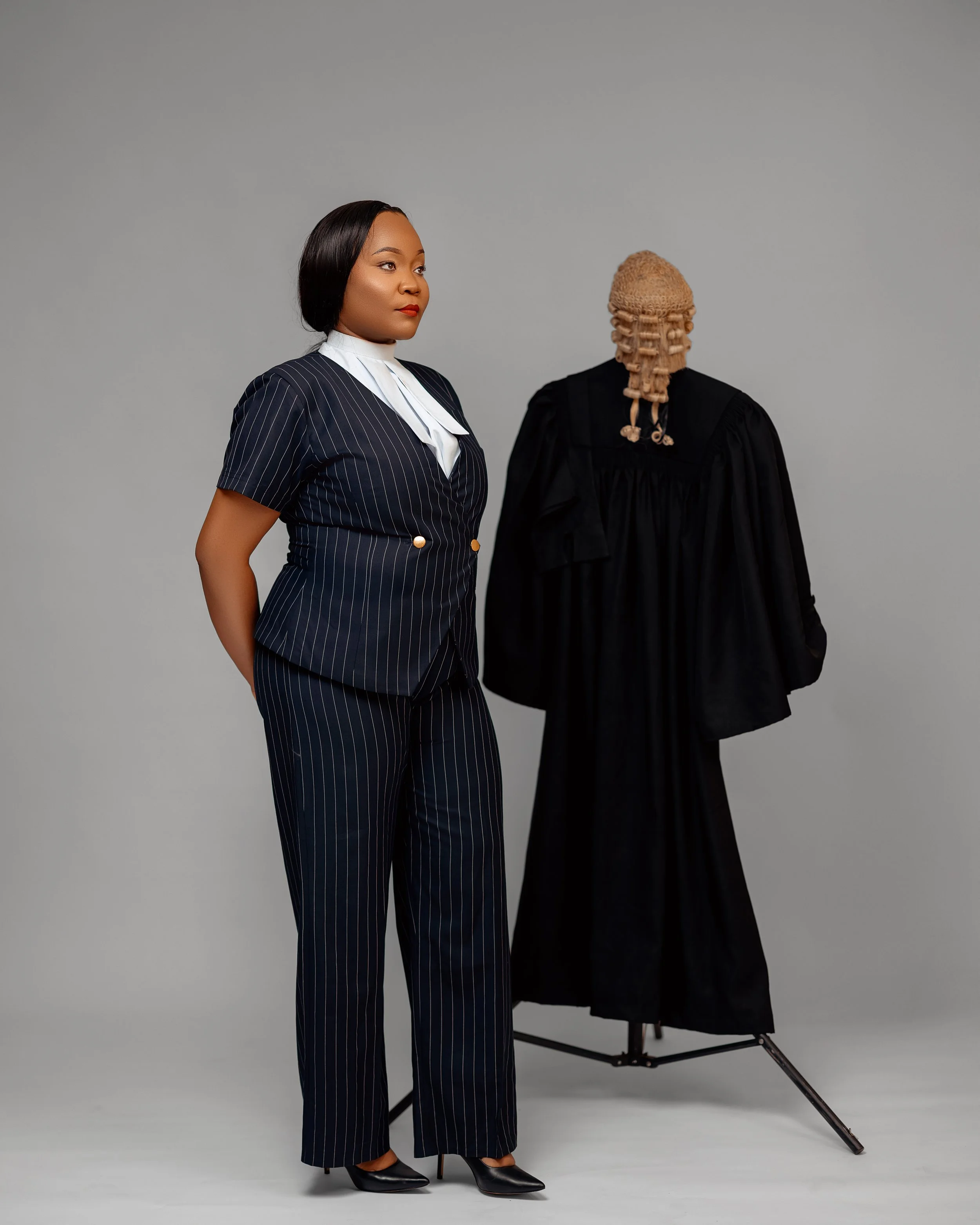A woman in a black and white pinstriped pantsuit standing next to a traditional African mask on a stand. The woman has short black hair and red lipstick.