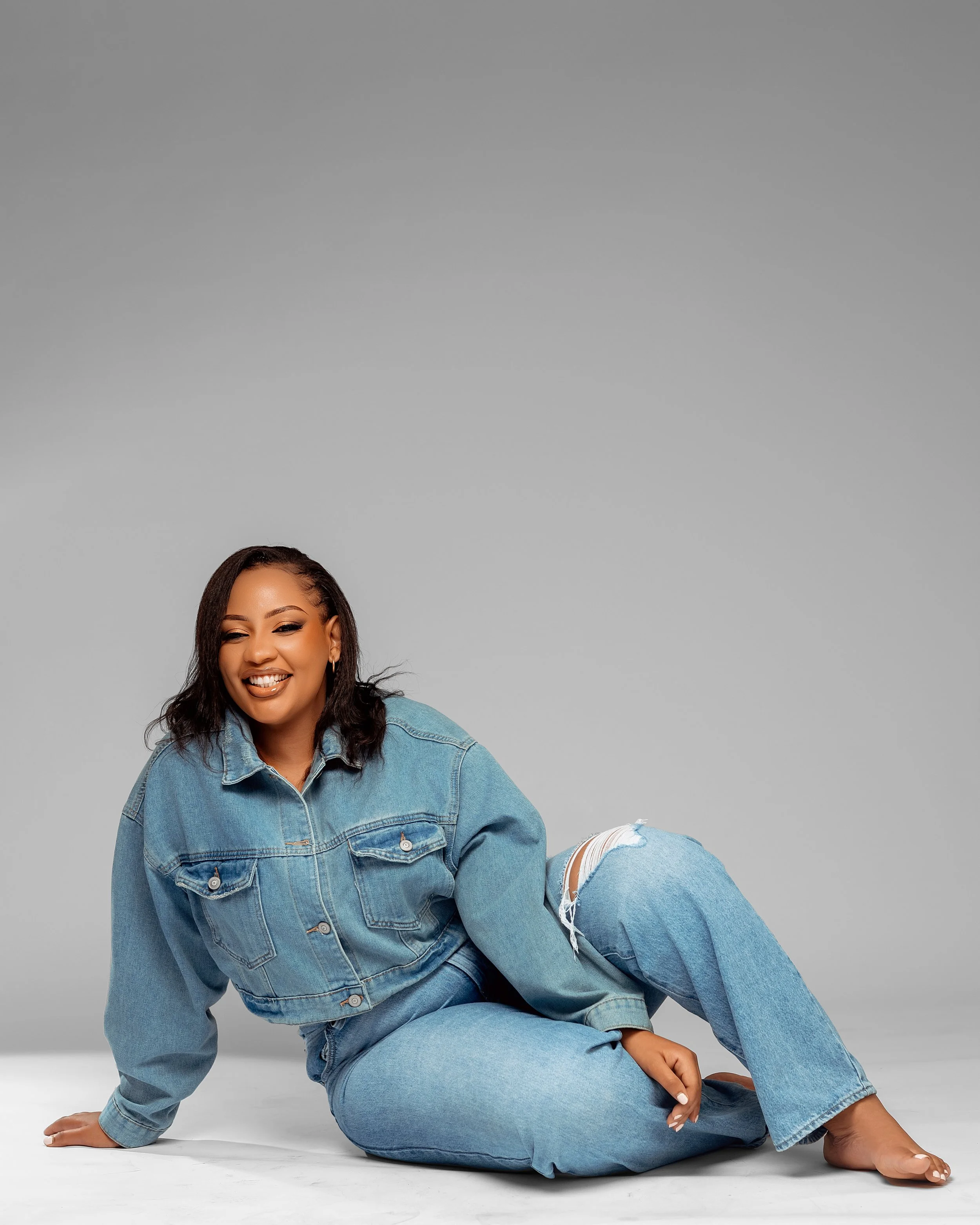 Woman wearing a denim jacket and ripped jeans, sitting on the floor against a plain gray background, smiling.