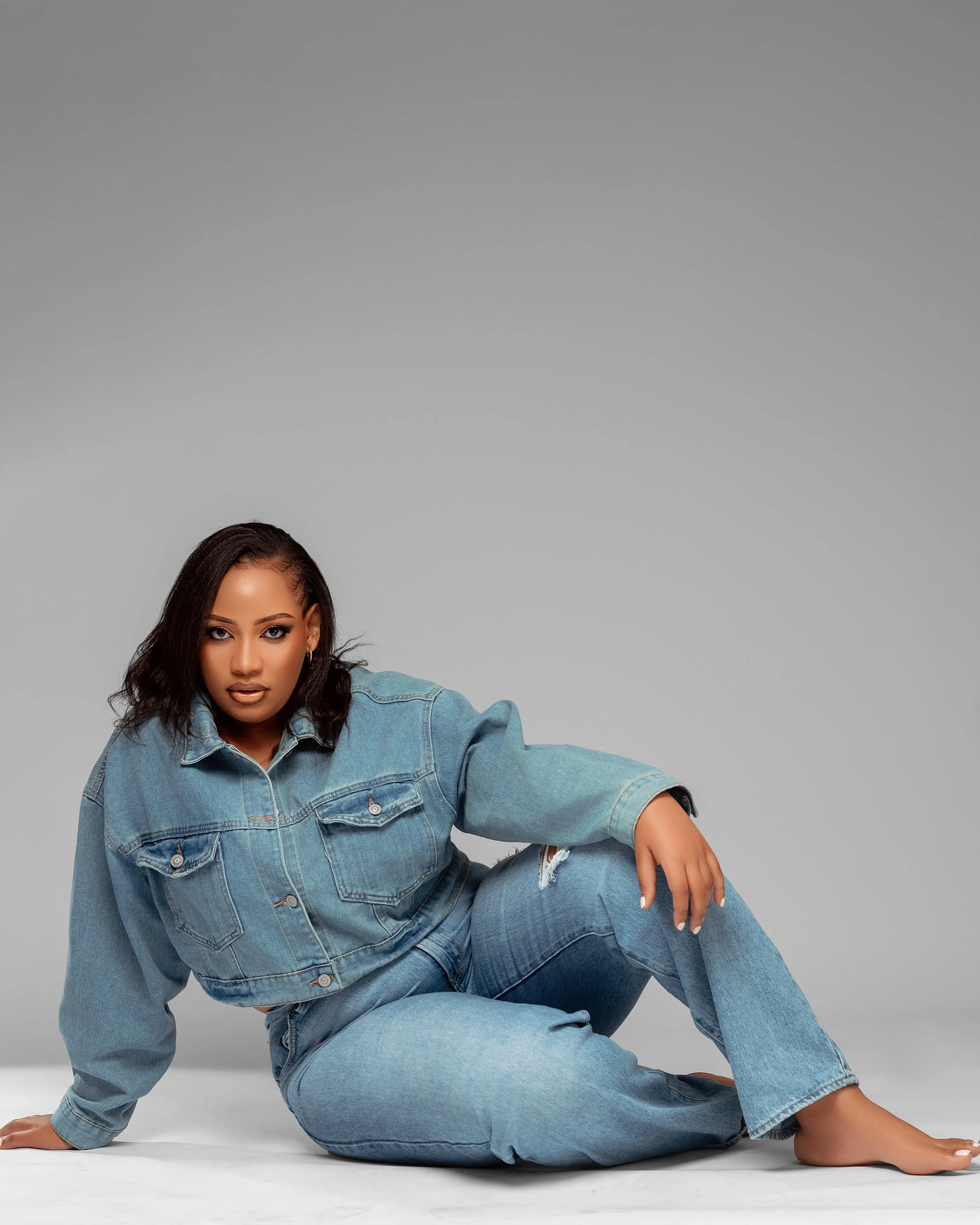 A woman in a denim jacket and jeans poses on a plain grey background.
