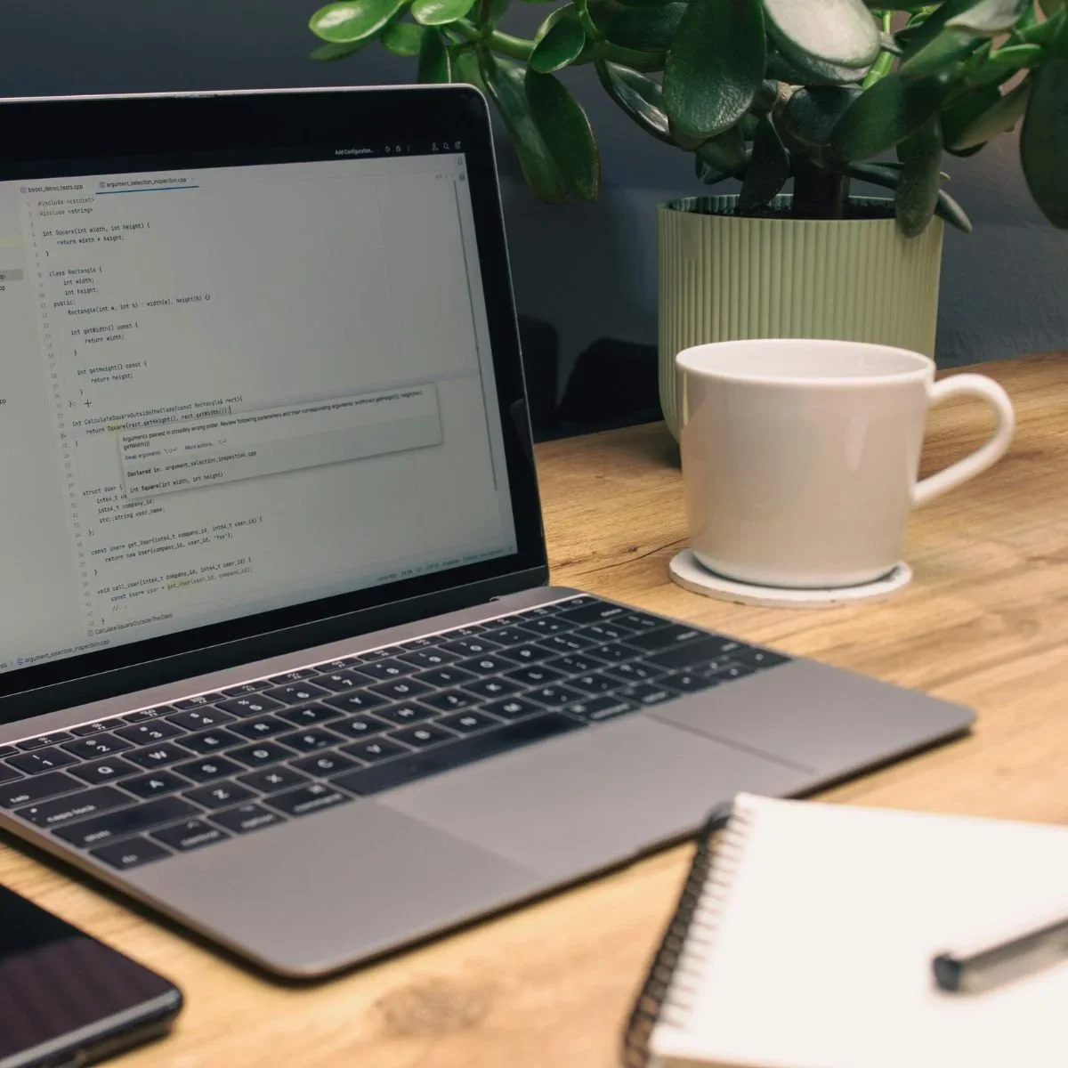 Affordable home office desk setup with open laptop displaying code, white coffee mug, spiral notebook, smartphone, and potted green plant on wood surface by HM Remodeling in South Bend, Indiana
