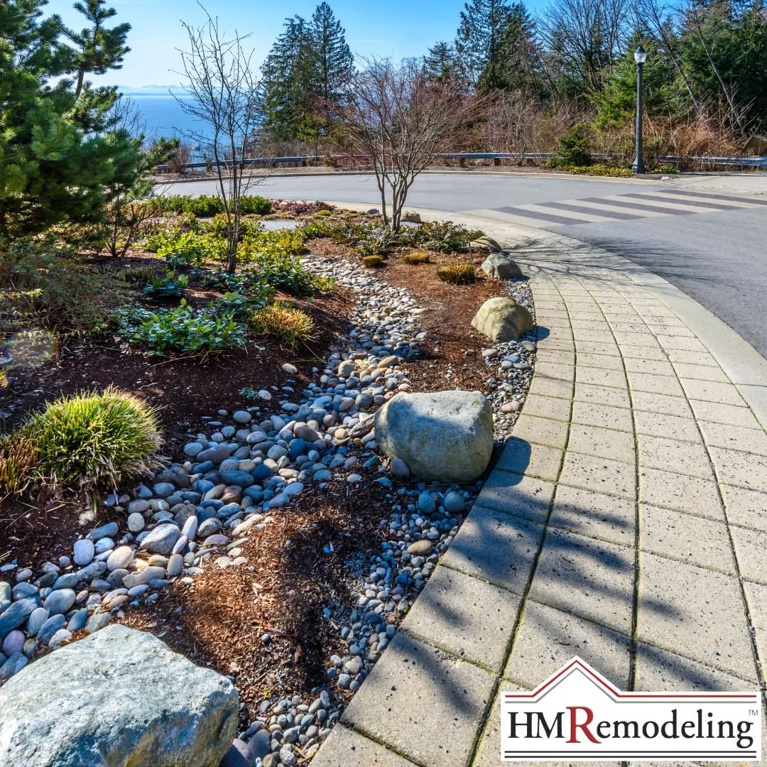 Paver driveway bordered by river rock drainage channel, mulched landscape bed with mixed shrubs and boulders, and evergreen plantings in a South Bend, Indiana home exterior improvement