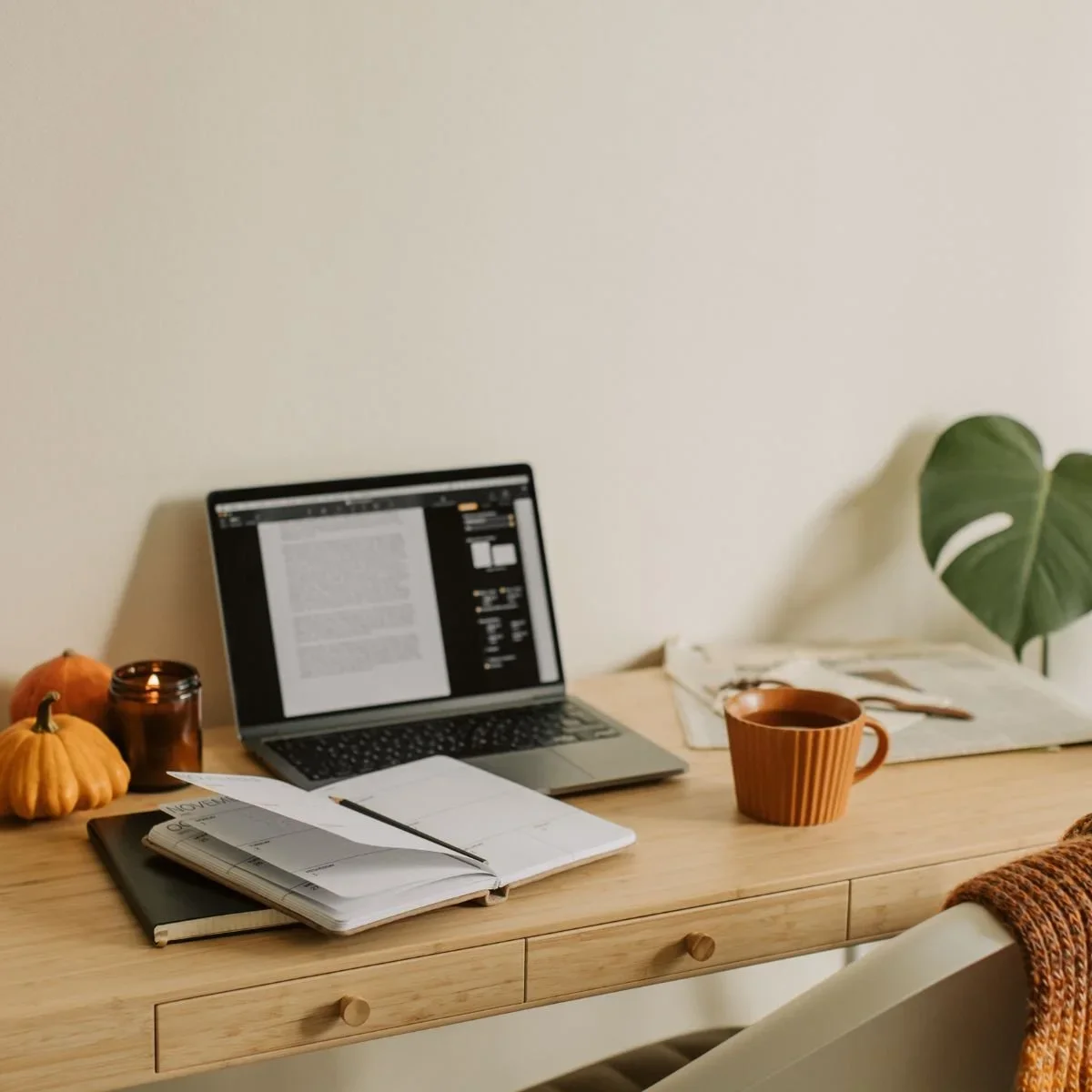 Cozy home office makeover with light wood desk, open planner, laptop, terracotta mug, mini pumpkins, candle, and monstera leaf plant against cream wall by HM Remodeling in South Bend, Indiana