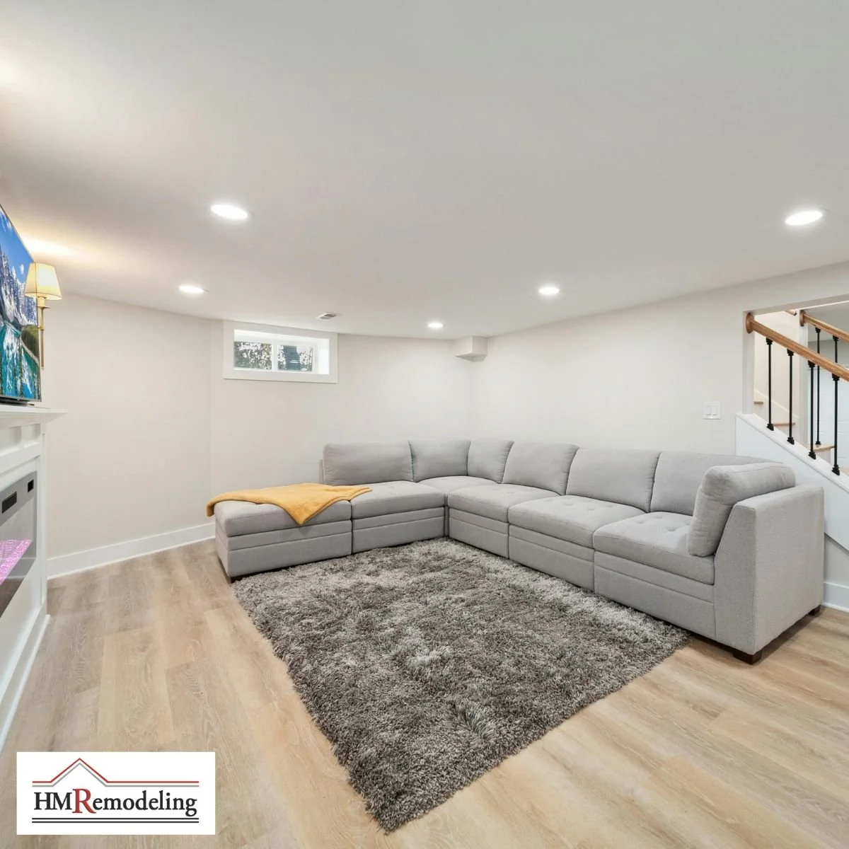 Finished basement living room with large gray sectional sofa, shag area rug, light LVP flooring, recessed lighting, egress window, white built-in cabinetry, and wood staircase with black iron railing by HM Remodeling in South Bend, Indiana