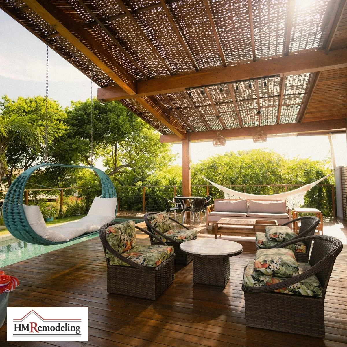 Covered outdoor patio deck with wood pergola and woven shade roof, hanging teal swing chair, wicker lounge seating with tropical cushions, hammock, and pool view by HM Remodeling in South Bend, Indiana