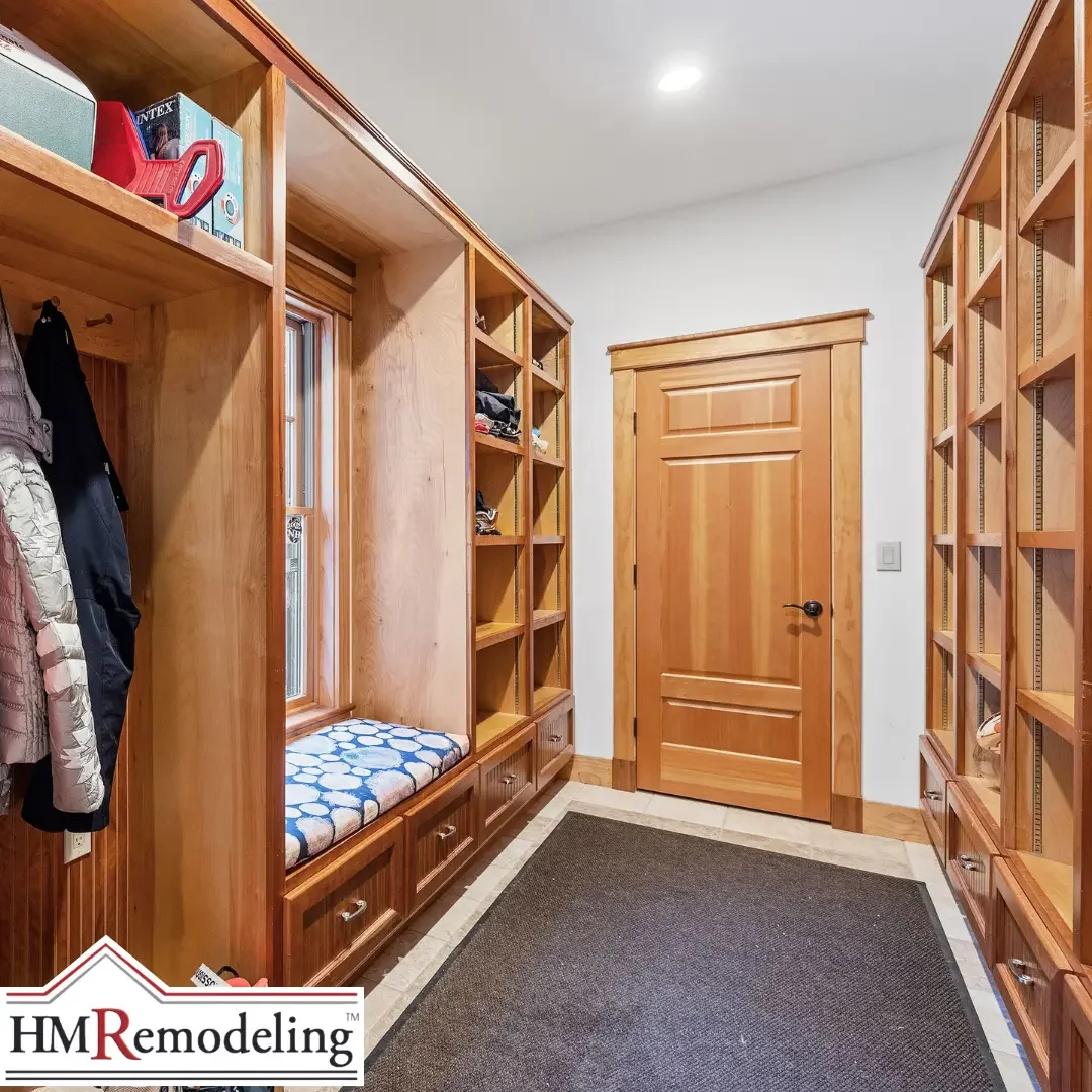 A spacious custom mudroom in a South Bend, Indiana home with floor-to-ceiling natural wood built-in shelving, a cushioned bench with drawers beneath, coat hooks on the left wall, and tile flooring with a dark area rug