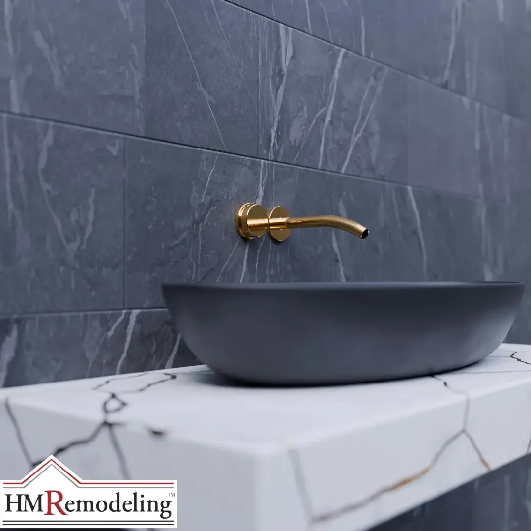 A dark gray vessel sink on a white marble-look counter with a wall-mounted brushed gold faucet against gray marble tile in a Michiana area bathroom.