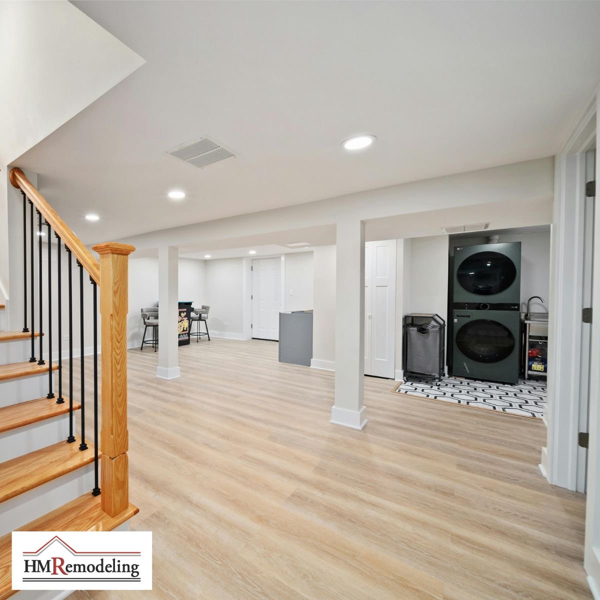 Finished basement with light LVP flooring, white painted walls, recessed lighting, white support columns, wood staircase with black iron balusters, and open laundry room with stacked washer and dryer by HM Remodeling in South Bend, Indiana