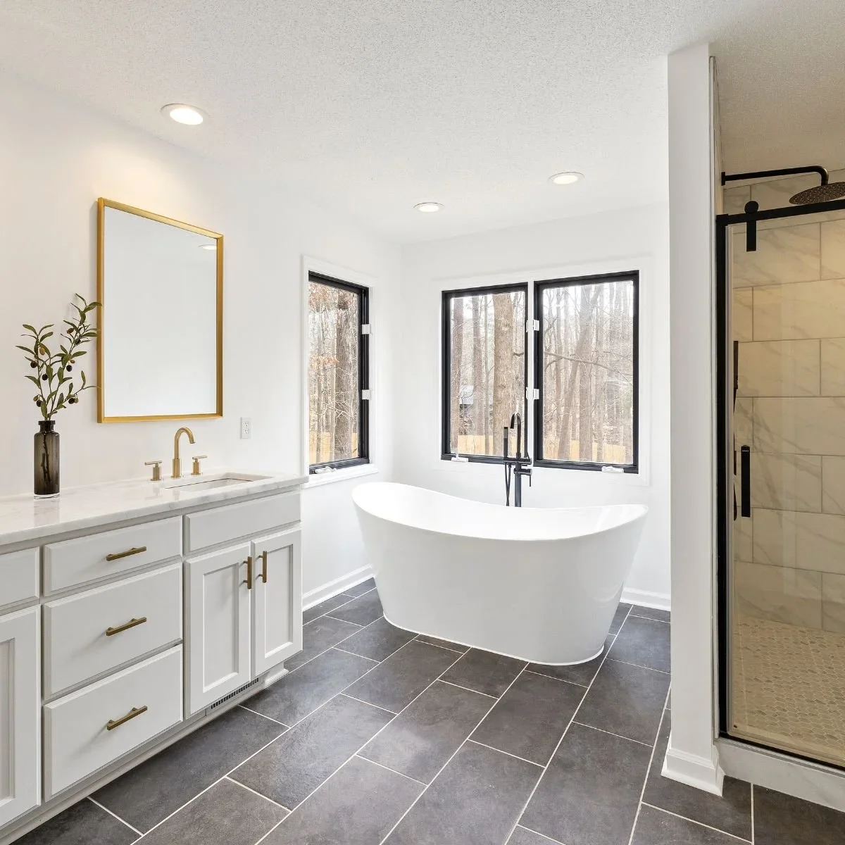 Winter bathroom remodel with freestanding soaking tub, white shaker vanity with gold hardware, dark gray tile flooring, black-framed windows, and walk-in shower with marble tile by HM Remodeling in South Bend, Indiana