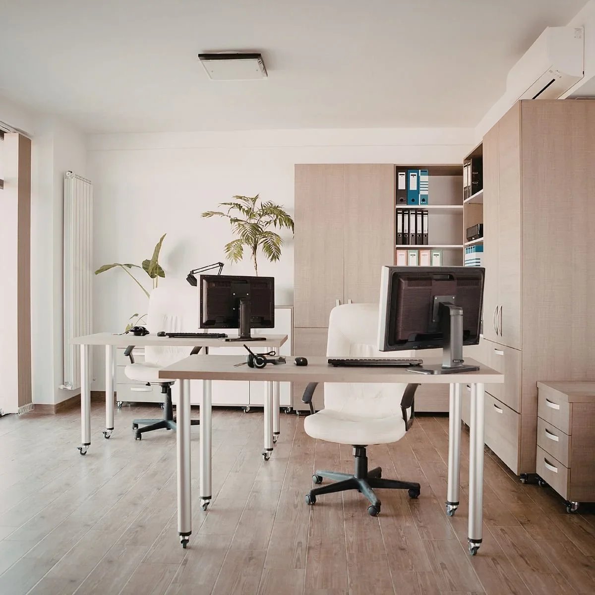 Modern home office makeover with dual workstation desks, white rolling chairs, floor-to-ceiling wood storage cabinets and shelving, hardwood flooring, and indoor plants by HM Remodeling in South Bend, Indiana